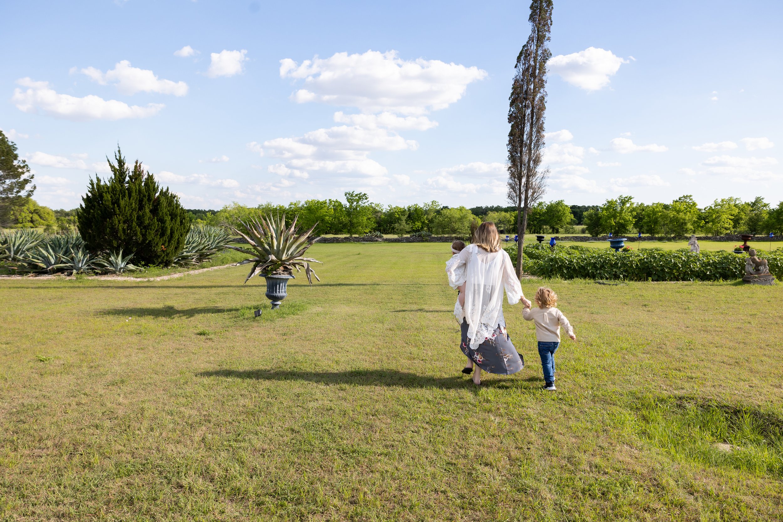 Outdoor lifestyle family portrait featuring parents and three young children sitting together in a grassy field with wildflowers and a stone wall backdrop. Captured in natural light, this candid family photo highlights connection, joy, and timeless s