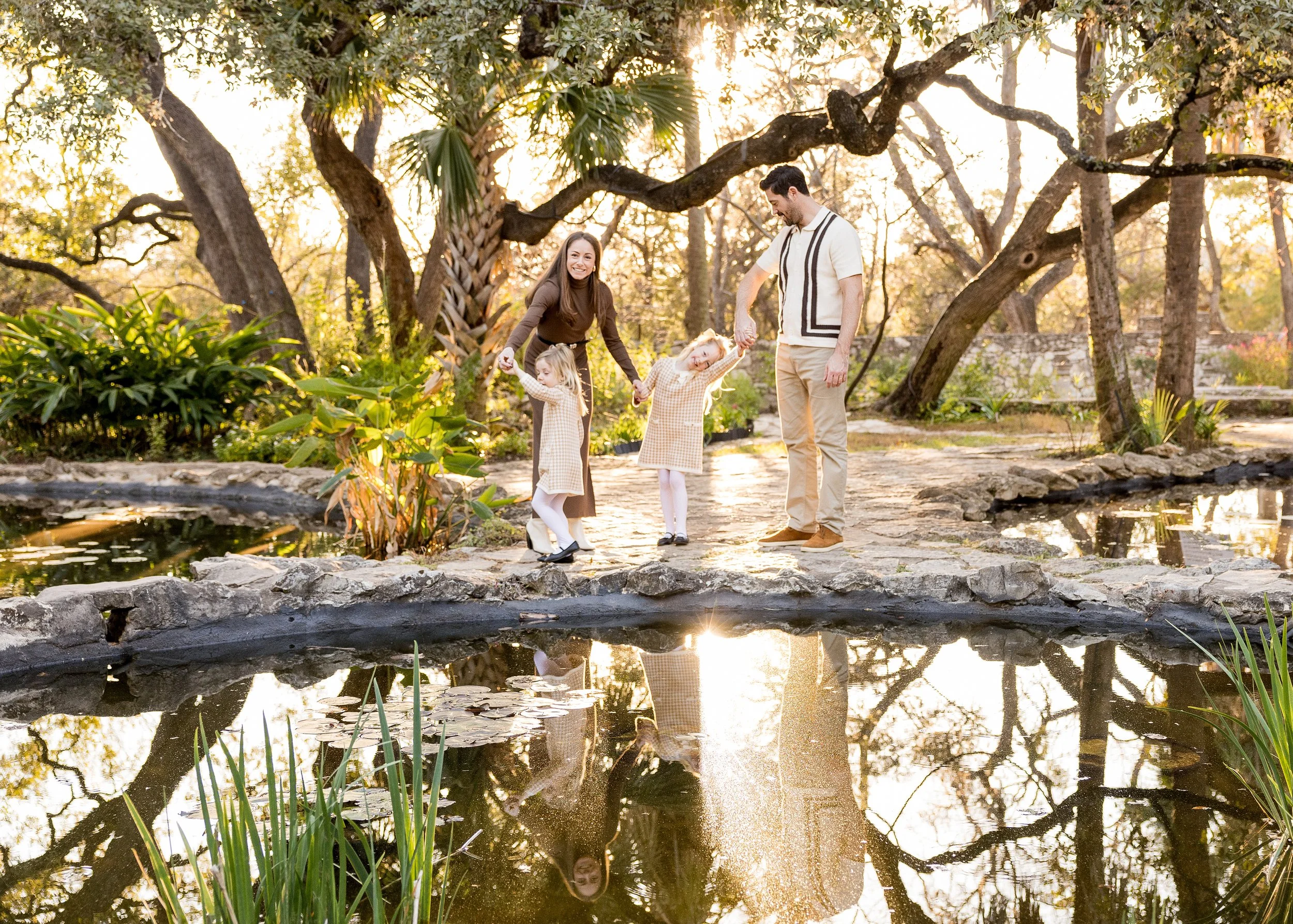 A candid family moment captured during golden hour as parents walk hand-in-hand with their two children along a tree-lined path. Soft natural light filters through the trees, creating a warm, timeless atmosphere that highlights connection, movement, 
