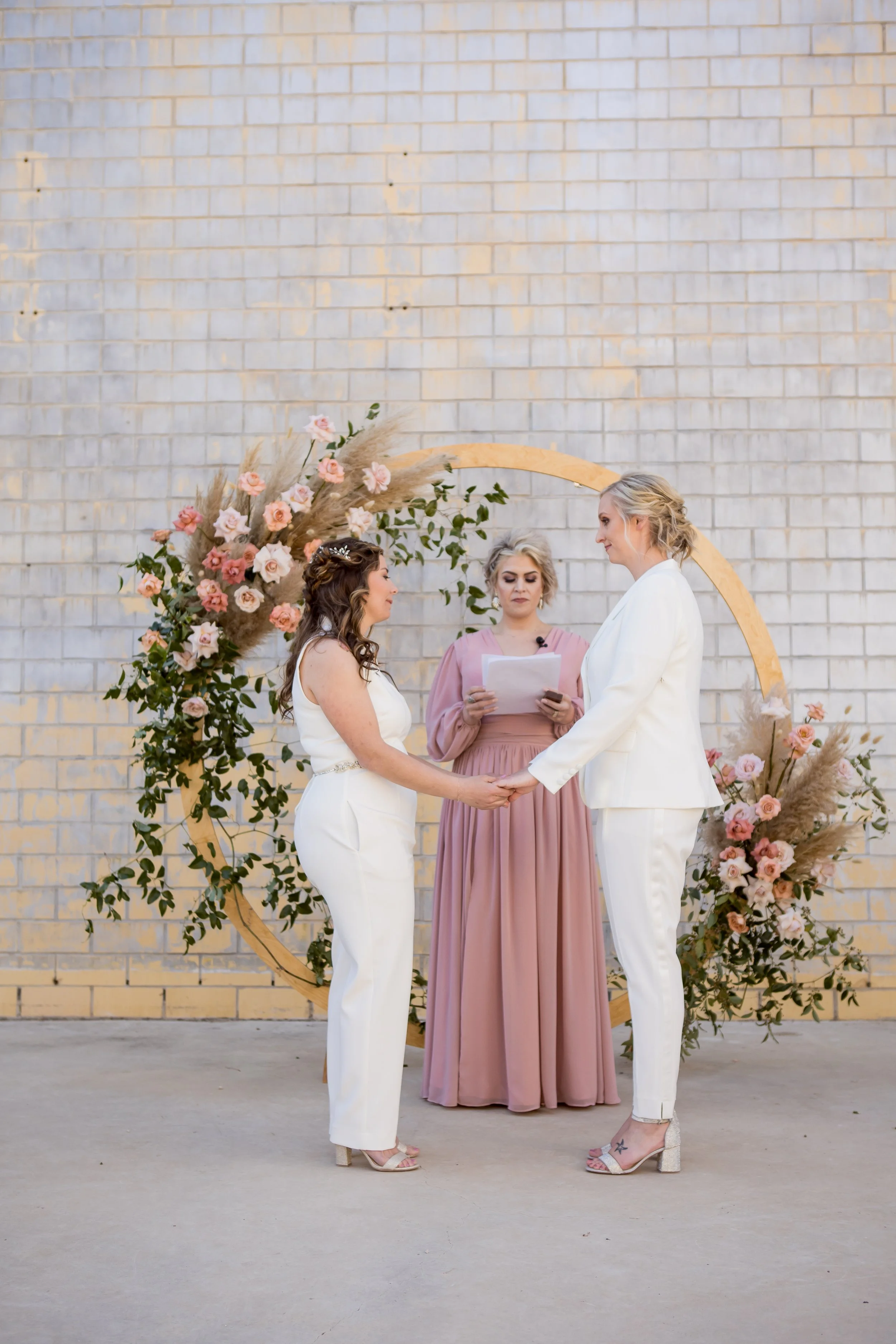 An intimate same-sex wedding ceremony featuring two brides exchanging vows beneath a modern circular floral arch.