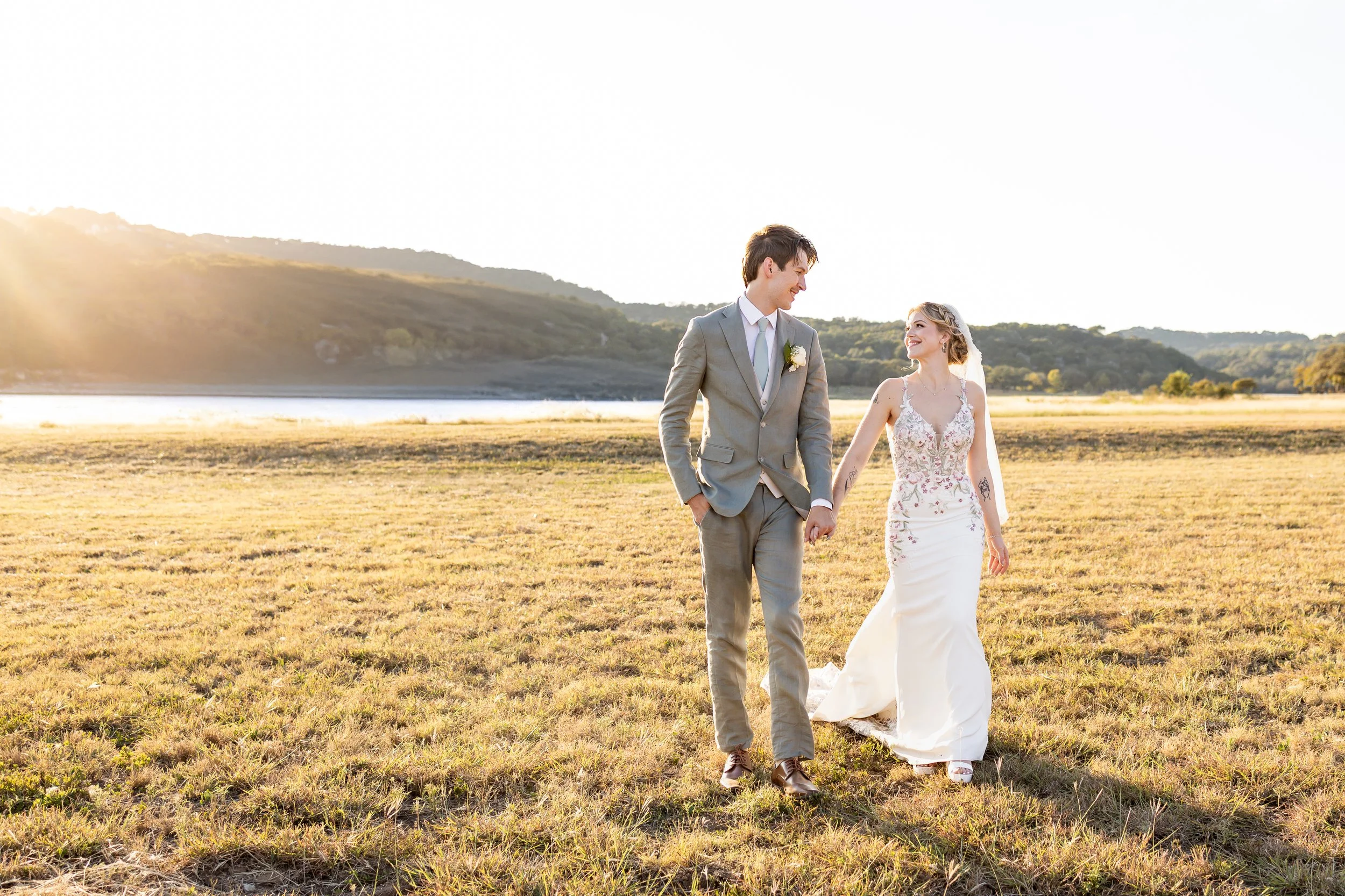 A bride and groom share a quiet, intimate moment at golden hour in an open field overlooking the water. Soft sunlight highlights the bride’s flowing gown and delicate details, creating a timeless, editorial wedding portrait that feels romantic, refin