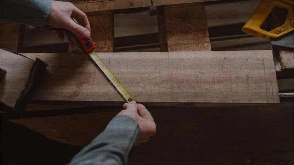 Person measuring a piece of wood with a tape measure on a workbench in a woodworking shop.