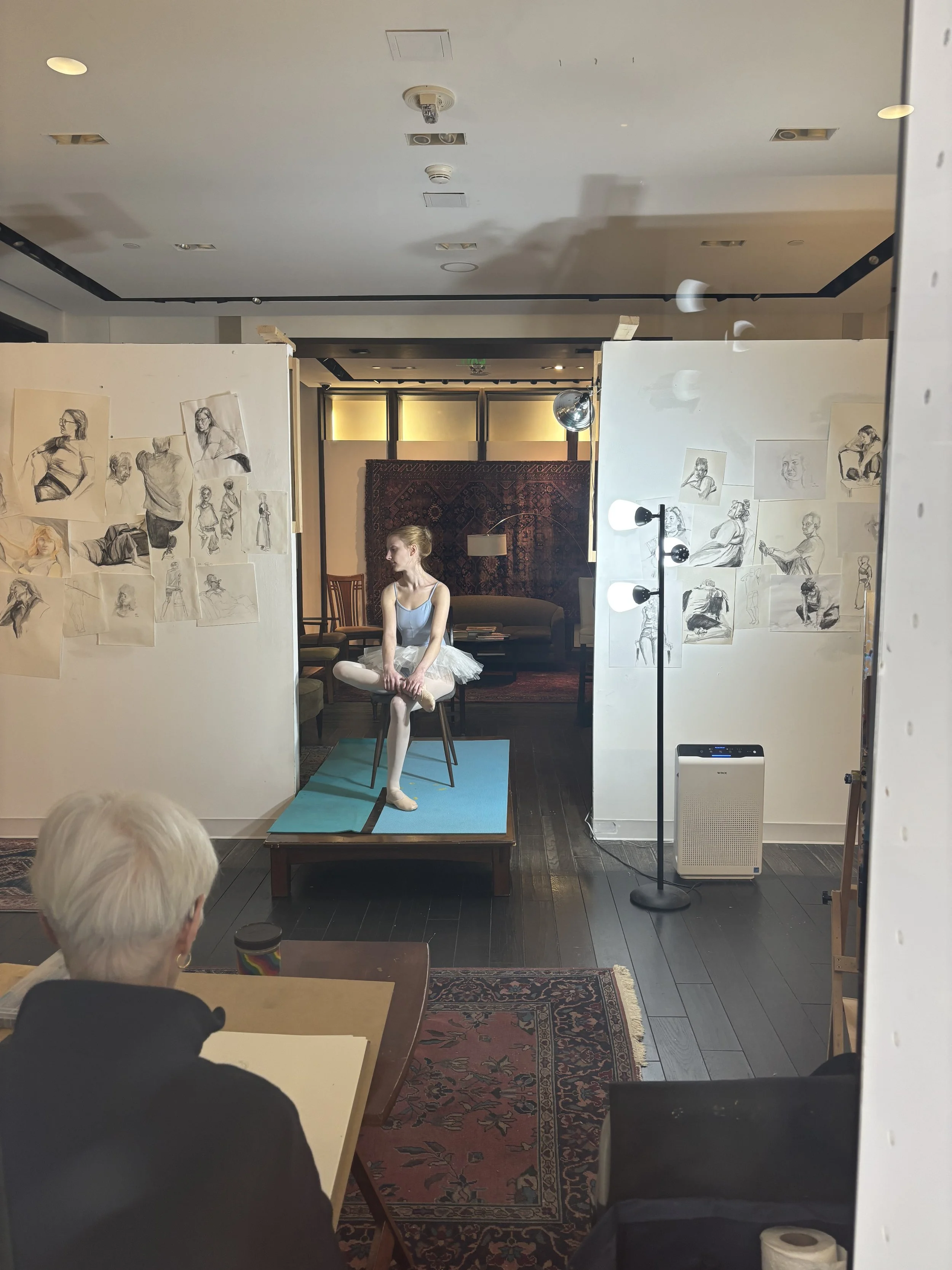 A ballet dancer sitting on a chair on a small platform in a studio, surrounded by sketches of various poses on display boards. A person with gray hair sits in the foreground, and the studio has a rug, chairs, and a decorative tapestry on the back wall.