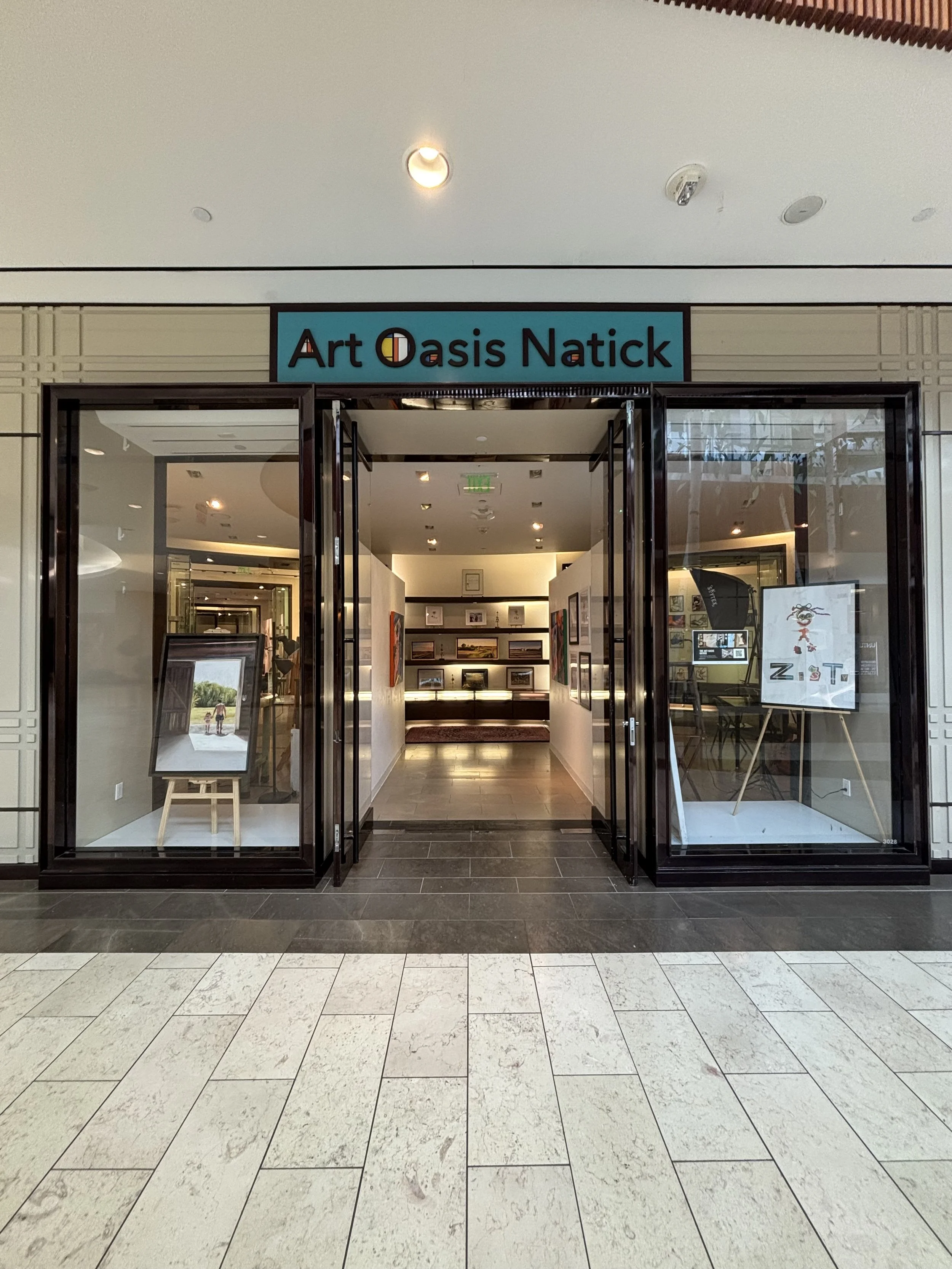 Entrance to an art gallery called Art Oasis Natick with glass doors, artworks inside, and tiled flooring outside.