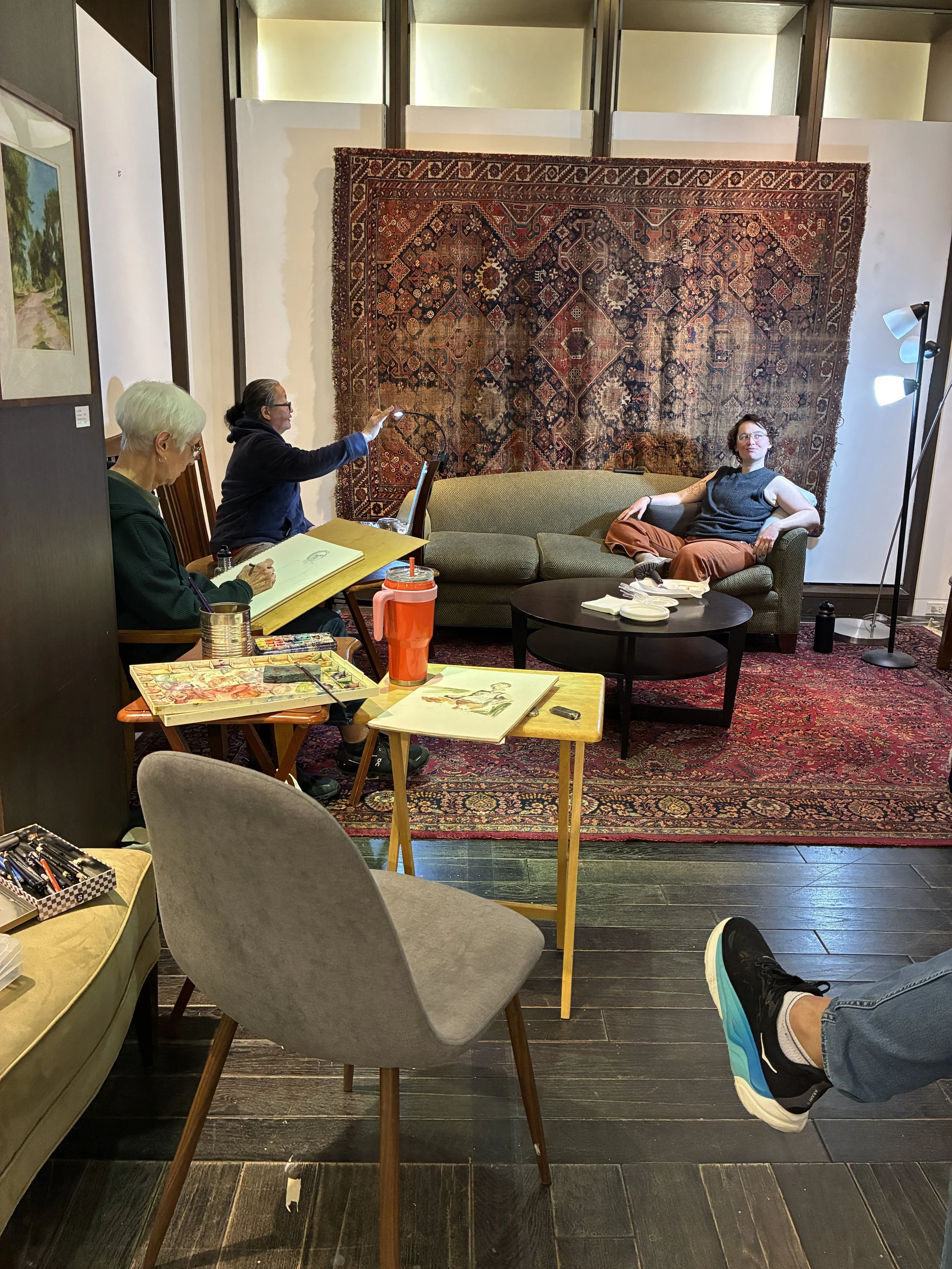 View from a chair showing a living room scene with a person reclining on a sofa, other people sitting at a table with painting supplies, and a large, intricate rug on the wall behind the sofa. Part of a person's shoe is visible at the bottom right.