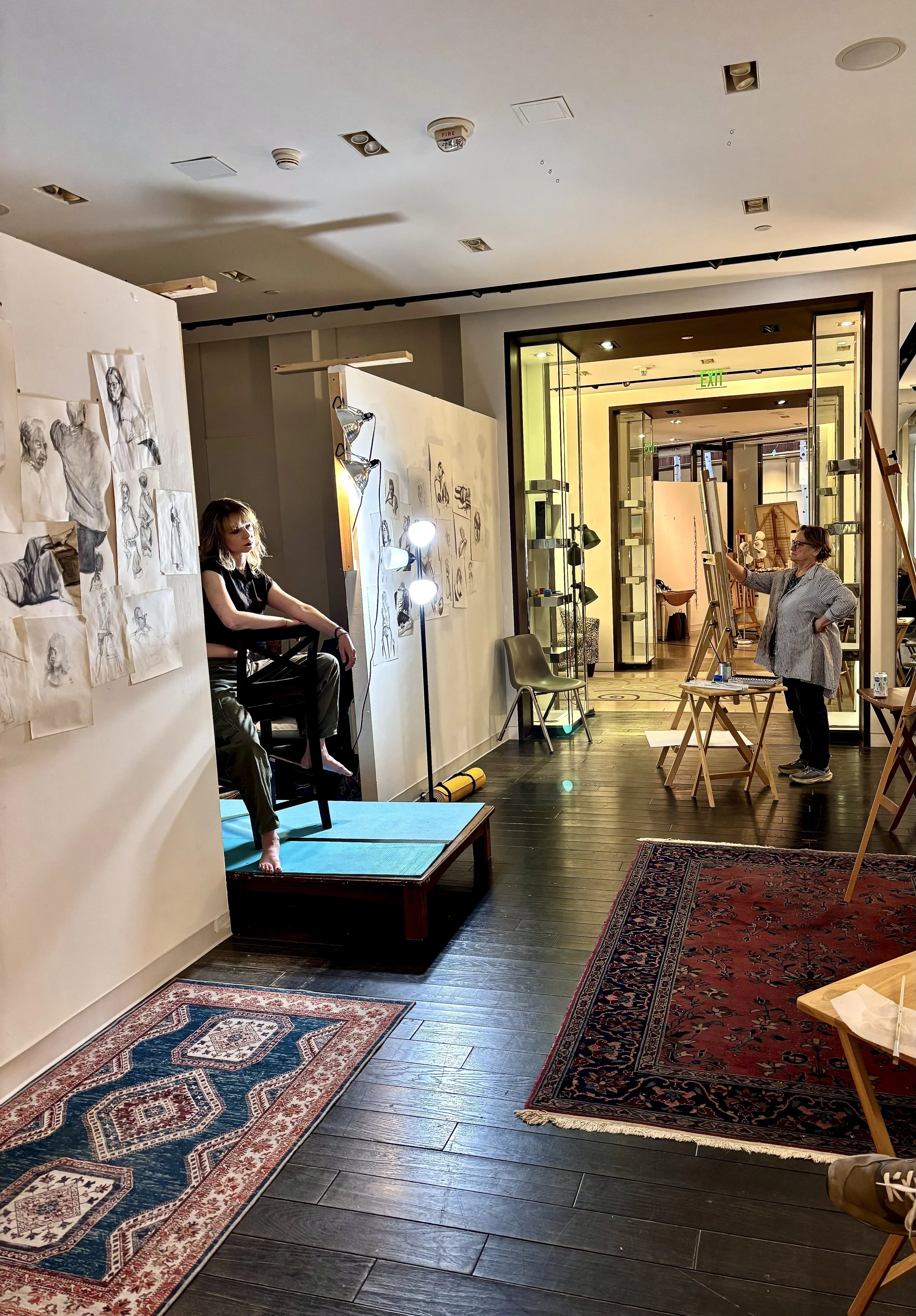 Art studio with drawings on the wall, a woman sitting on a chair, a woman standing and drawing, area filled with art supplies and rugs on wooden floor.