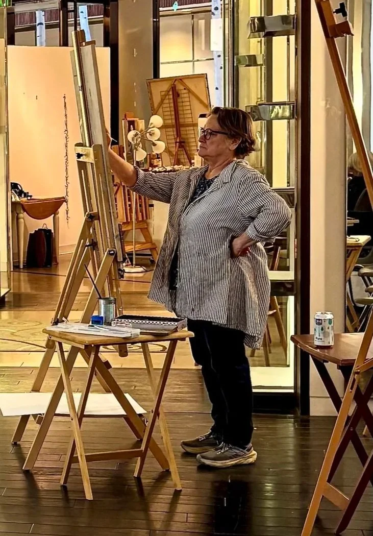 Woman wearing glasses and a striped jacket painting on a canvas in an art studio, with art supplies and furniture around her.