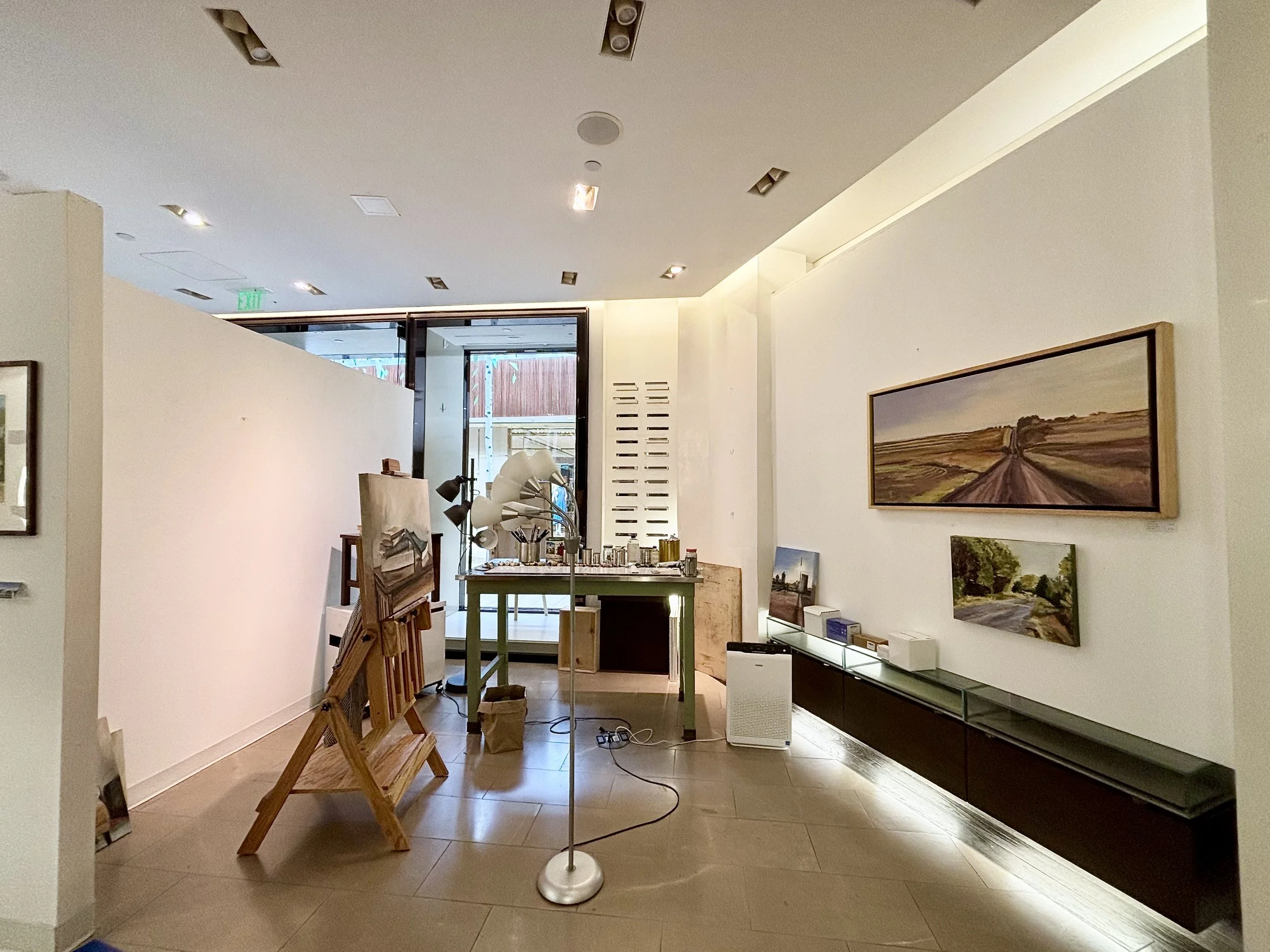 Interior view of an art gallery with artworks on the walls, a table with art supplies, and lamps on a stand in the foreground.