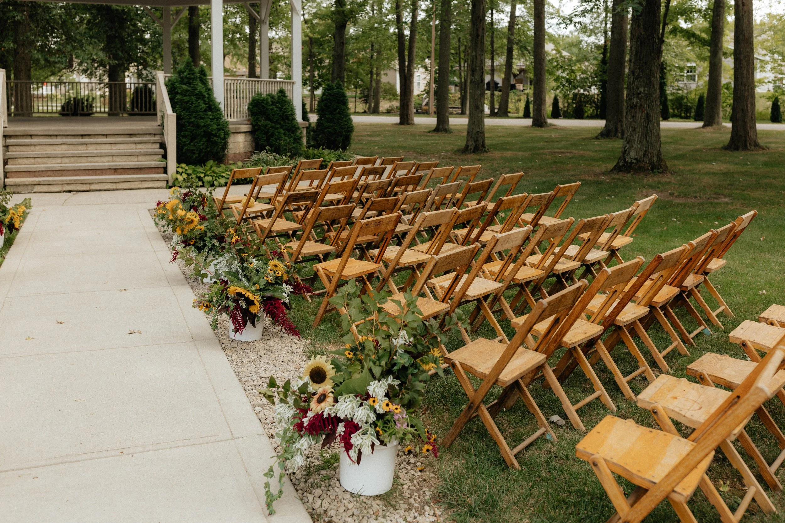 Outdoor wedding seating setup with wooden chairs arranged in rows on a lawn, decorated with flower arrangements in white pots along a paved aisle, with trees and a porch in the background.
