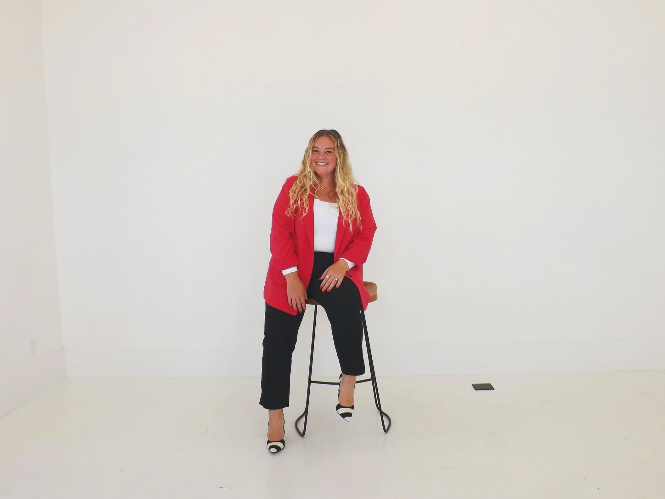 A woman with long curly blonde hair, wearing a red blazer, white top, black pants, and black-and-white striped shoes, is sitting on a brown stool in a white room with a minimalist background.