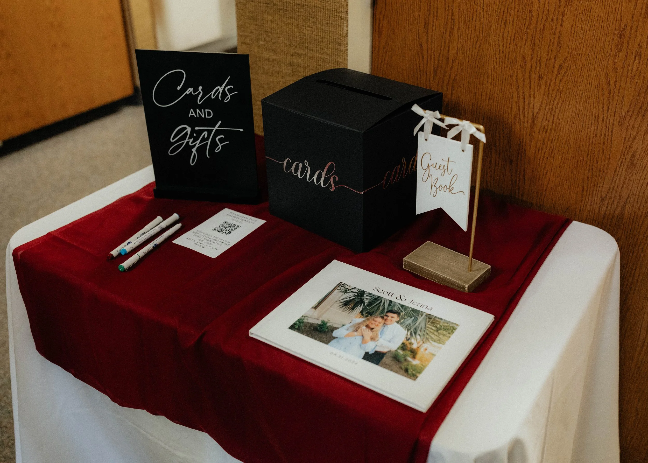 Wedding guestbook table with a photo album, a black gift box labeled 'cards,' a sign reading 'Cards and gifts,' a white sign labeled 'Guest Book' on a stand with ribbons, three pens, and a QR code paper, all on a red tablecloth.