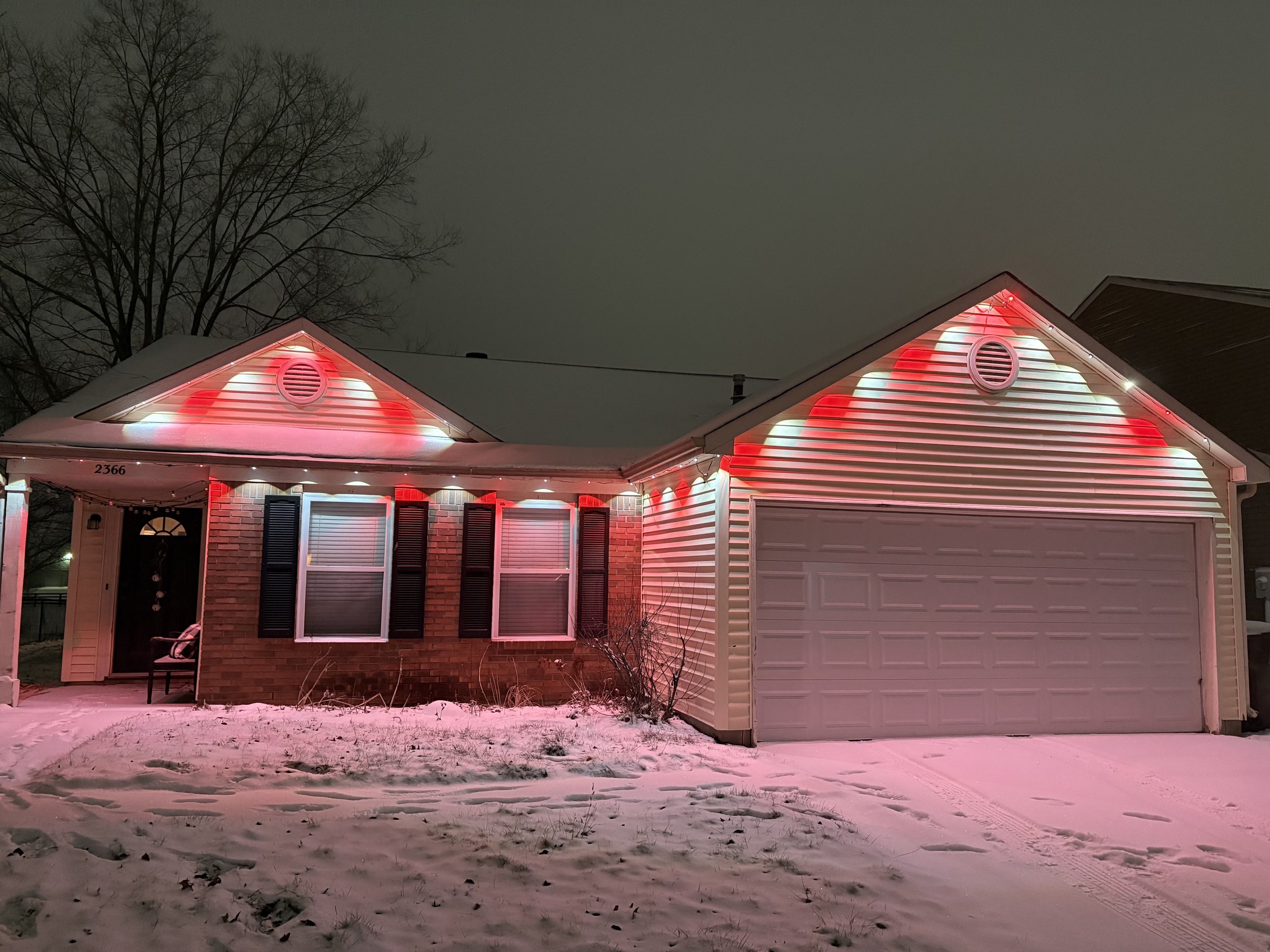 A house decorated with red and white Christmas lights, with snow on the ground and a leafless tree in the background at night.