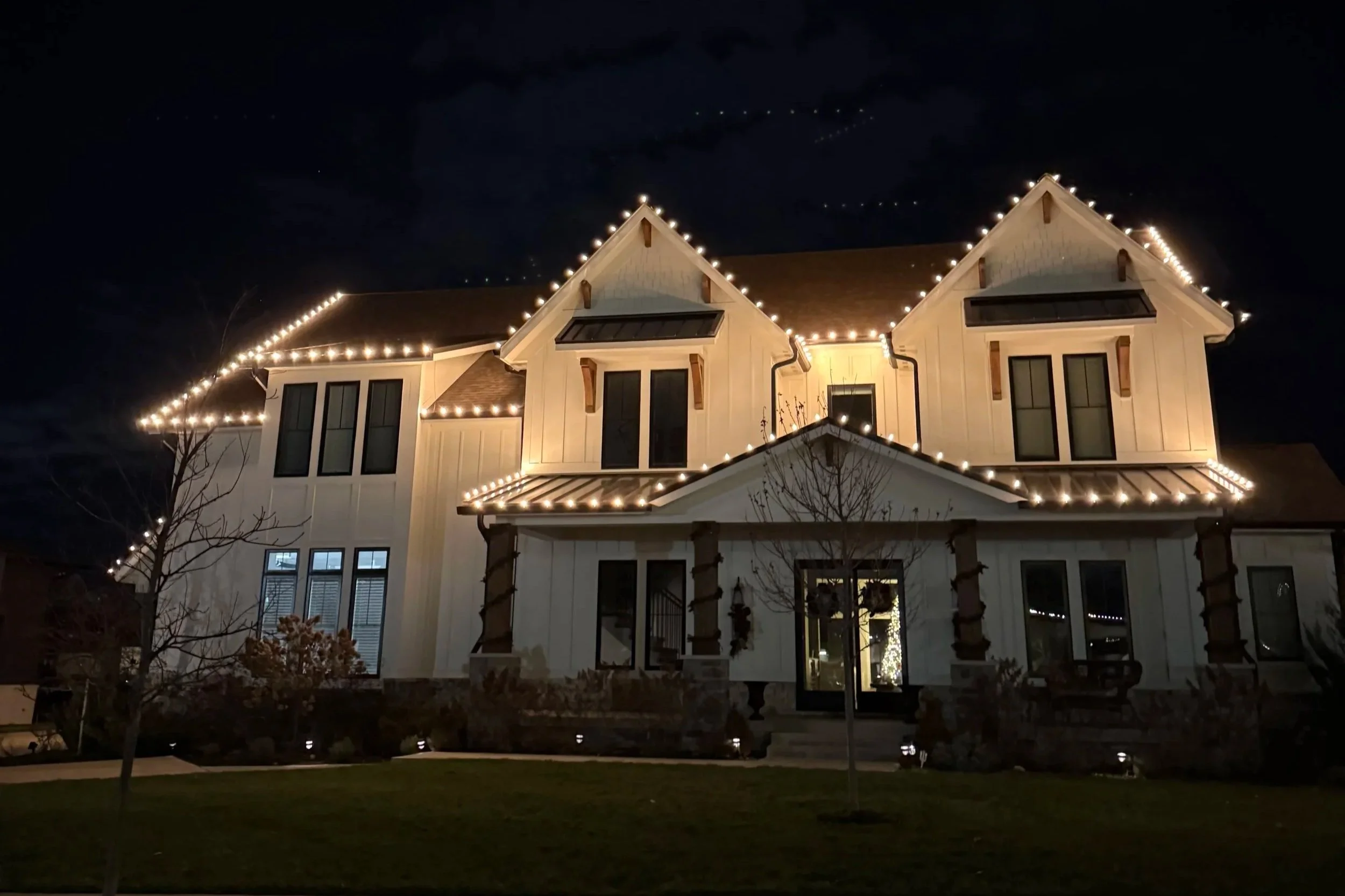 Night scene of a multi-story house decorated with string lights along the roof edges and in the front yard.
