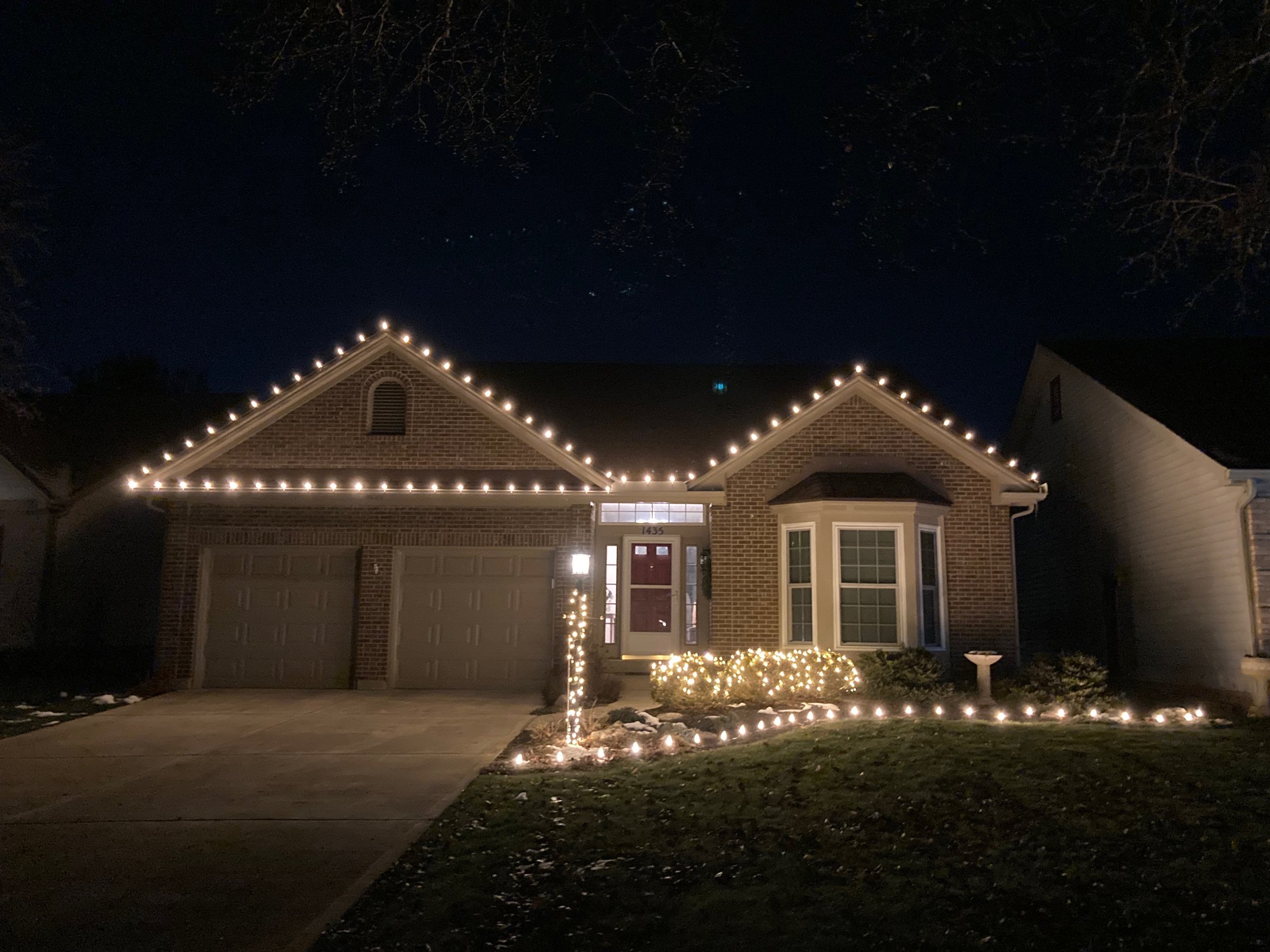 A suburban house decorated with white Christmas lights outlining the roof, windows, front door, and driveway, nighttime scene with dark sky and surrounding trees.