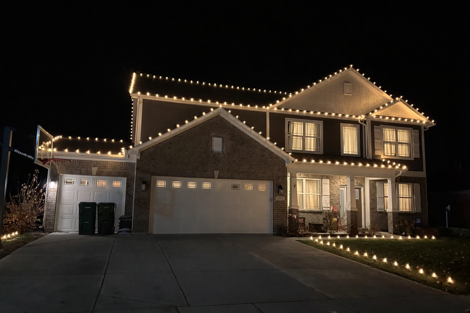 A two-story house decorated with string lights outlining the roof and yard at night. The house has a brick facade and illuminated windows.