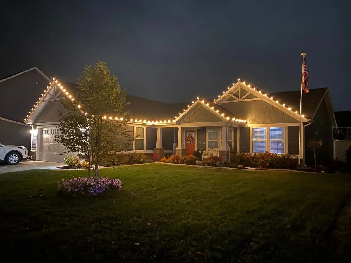 A house decorated with string lights along the roofline, with a front yard featuring a tree, flowers, a lawn, and an American flag on a flagpole, taken during nighttime.