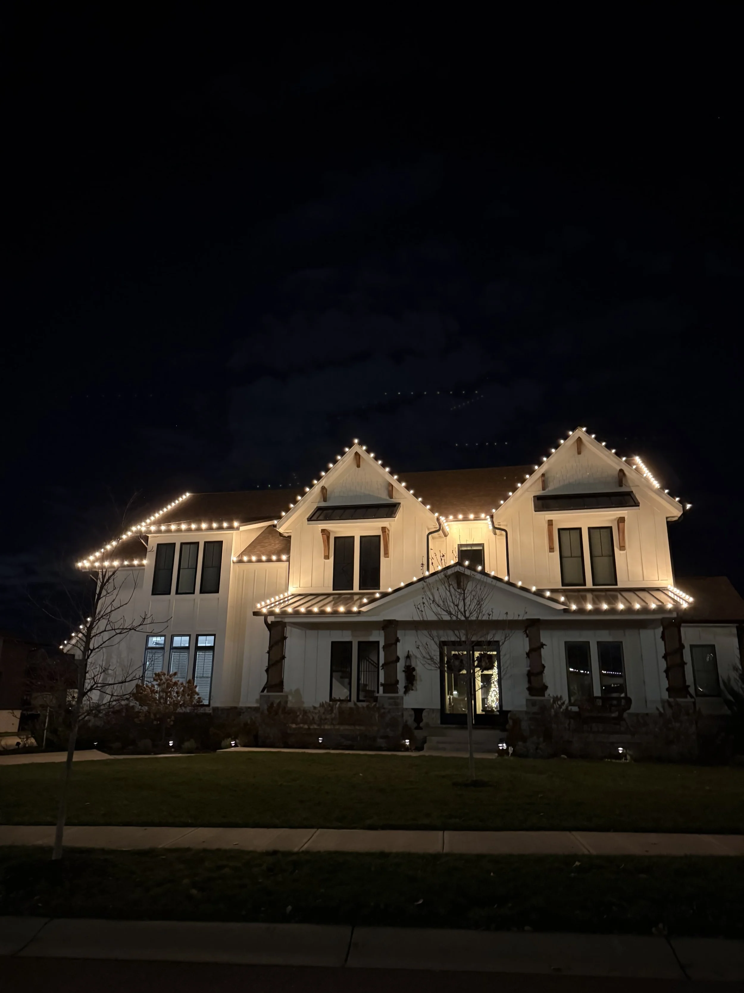 A large, multi-story house decorated with white string lights outlining the roof, windows, and porch area, set at night.