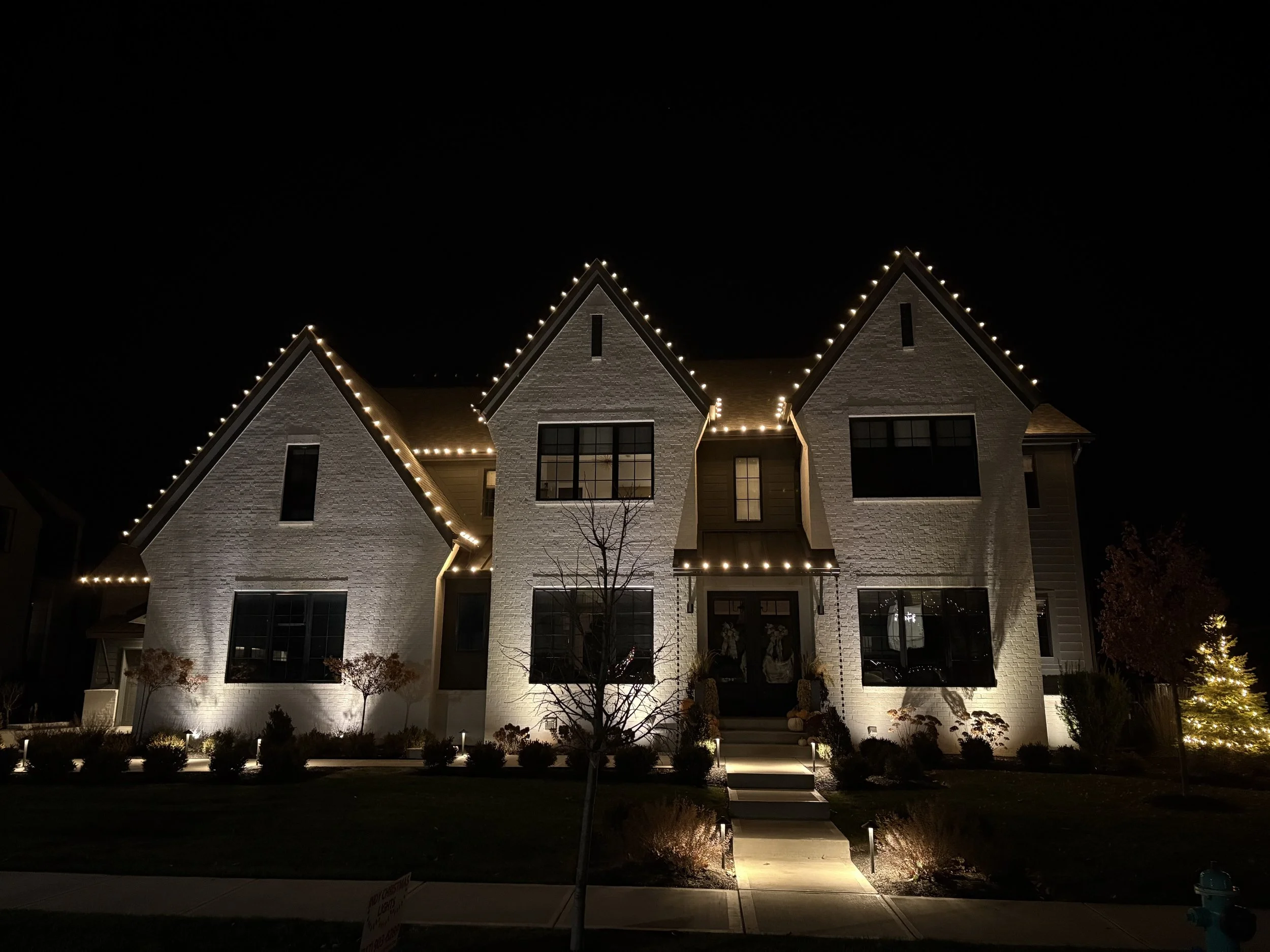 A modern house with triangular gabled roofs illuminated with string lights at night, with landscaped front yard and pathway.