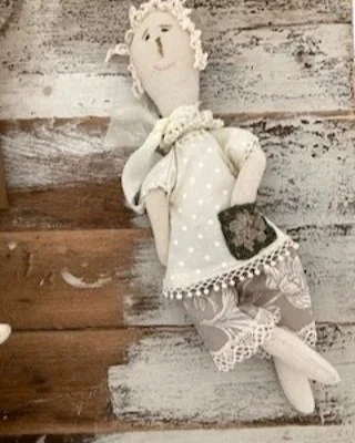 A handmade fabric doll with curly white hair, wearing a polka dot top, lace shorts, and holding a small black bag, lying on a rustic wooden surface.