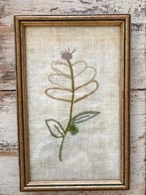 Framed embroidered artwork of a stylized plant with a small flower at the top, on a light fabric background.