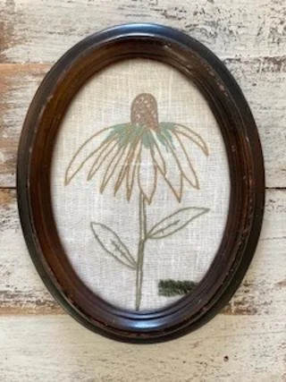 Framed embroidered botanical illustration of a flower with a large yellow and brown center and long green petals, on a light background.