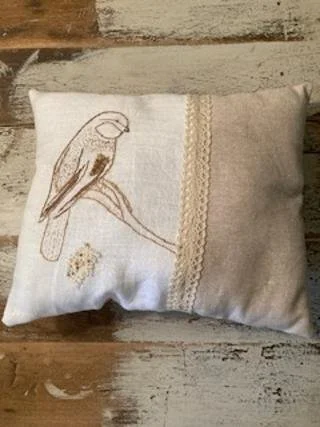 Decorative pillow with embroidered bird and lace detail on a rustic wooden surface.