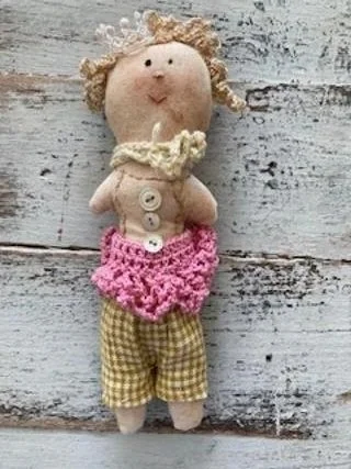 Folk art doll with curly yarn hair, wearing yellow plaid shorts and a pink crocheted skirt, standing against a weathered wooden background.