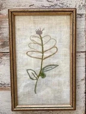 Framed embroidery of a simple flower with a green stem, two green leaves, and a yellow bloom on a light textured background.