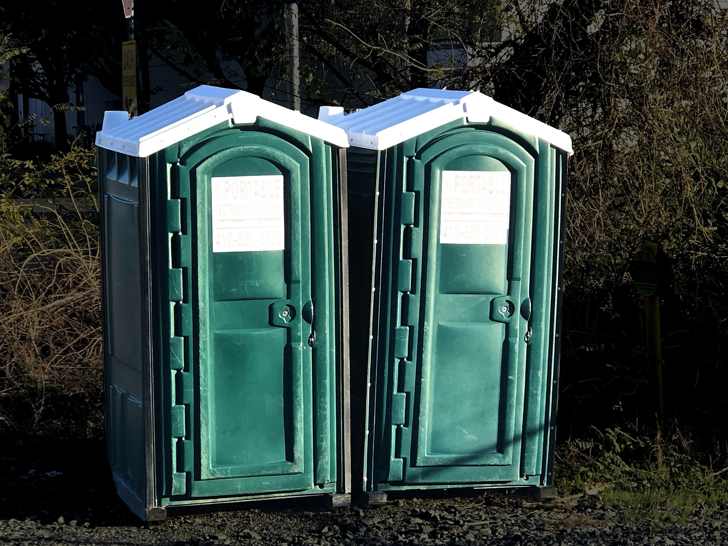 Two green portable toilets with white roofs standing side by side on gravel ground in an outdoor area with trees and bushes in the background.