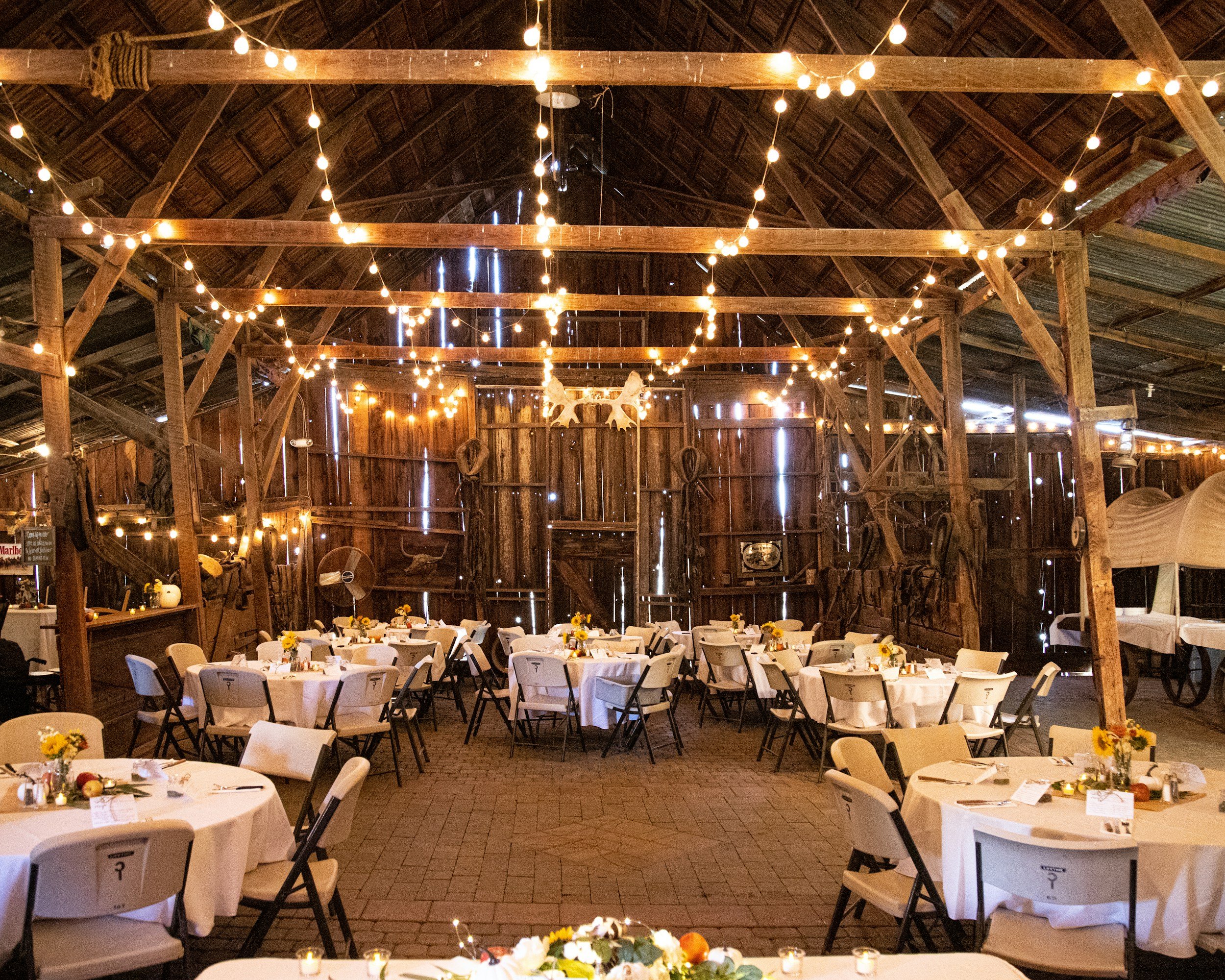 Indoor rustic barn setup with round tables covered in white tablecloths, decorated with small floral centerpieces, in a large wooden barn with string lights hanging from the ceiling, and horseshoe decorations on the back wall.