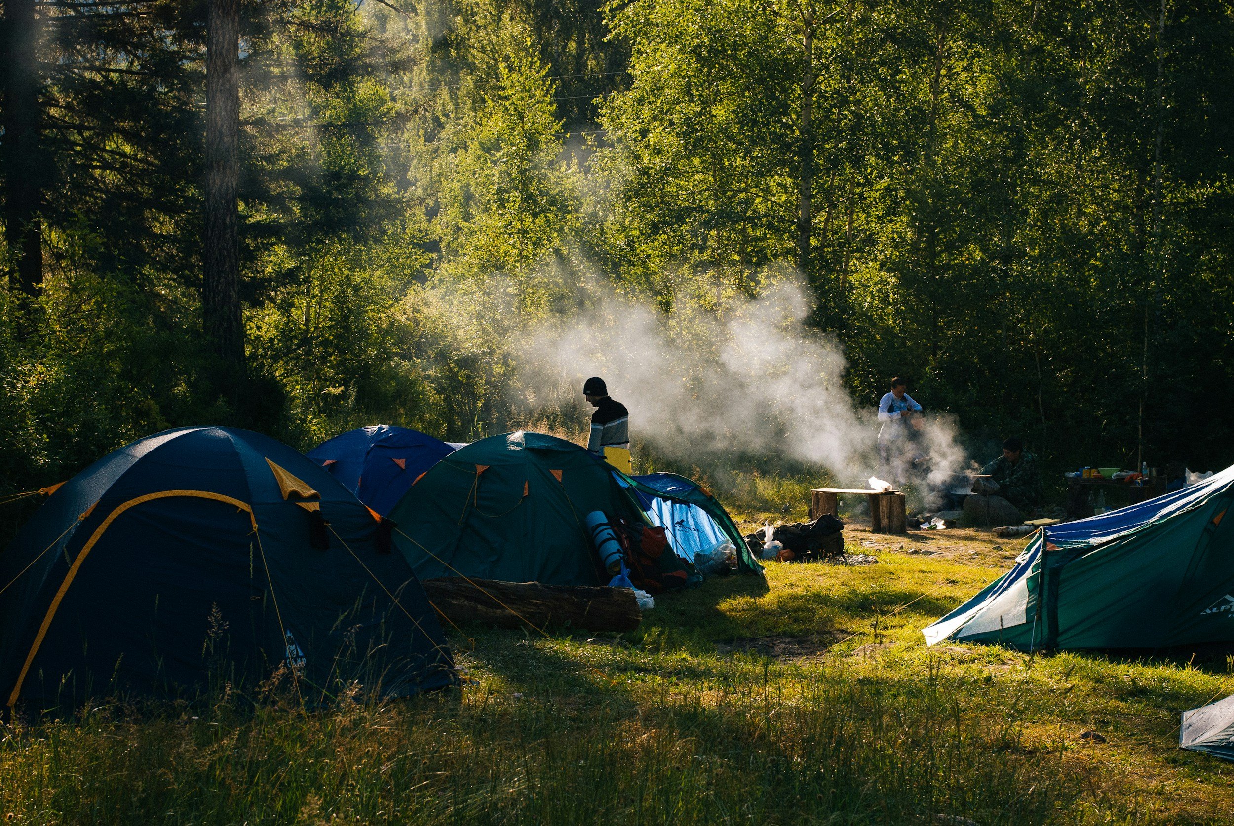 A group of people camping in a forest with multiple tents, some people standing near a campfire with smoke rising, and others sitting around it, surrounded by green trees and sunlight.