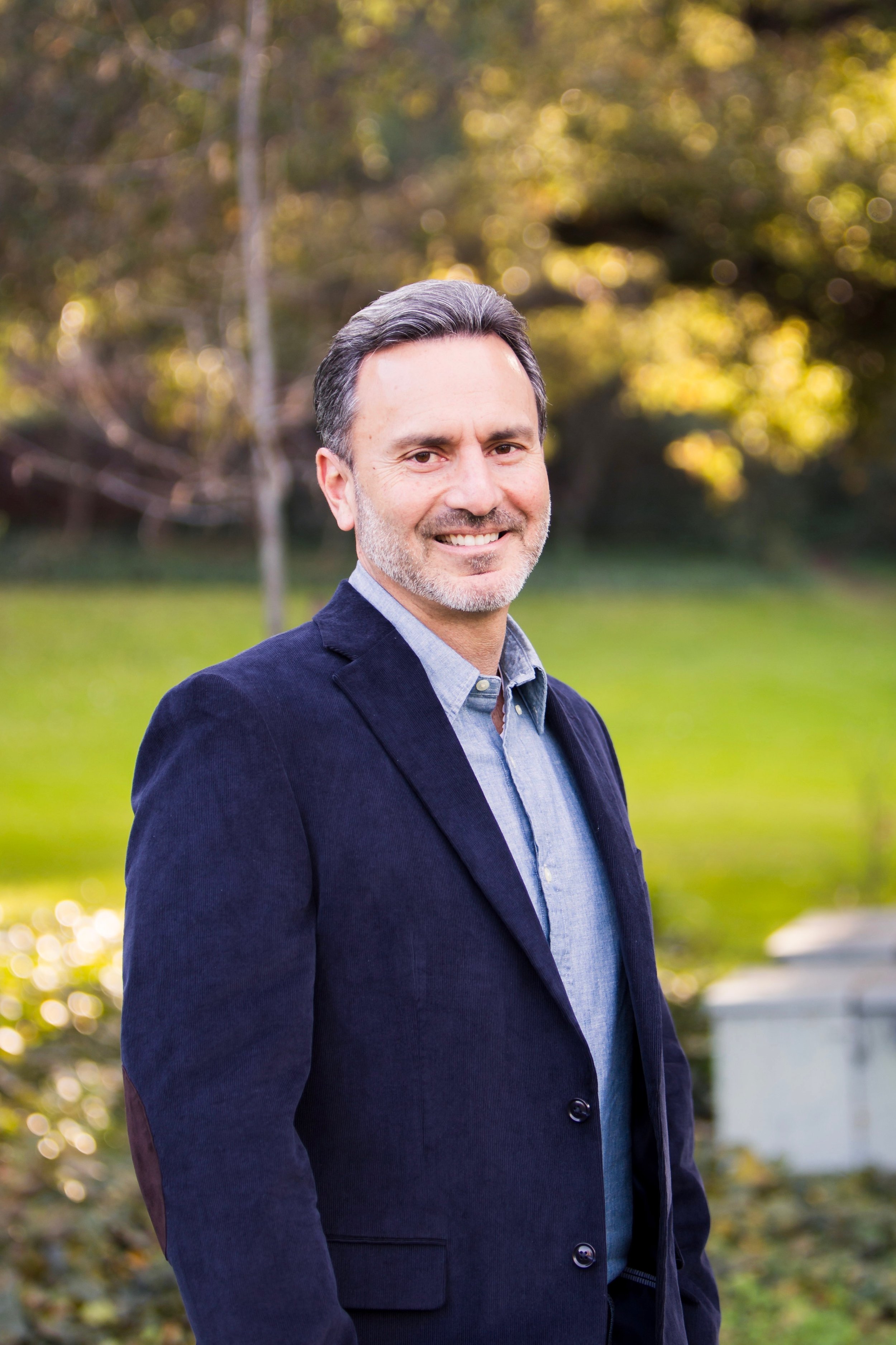 A smiling middle-aged man with dark hair and a beard, wearing a blue blazer and light blue shirt, standing outdoors with green grass and trees in the background.