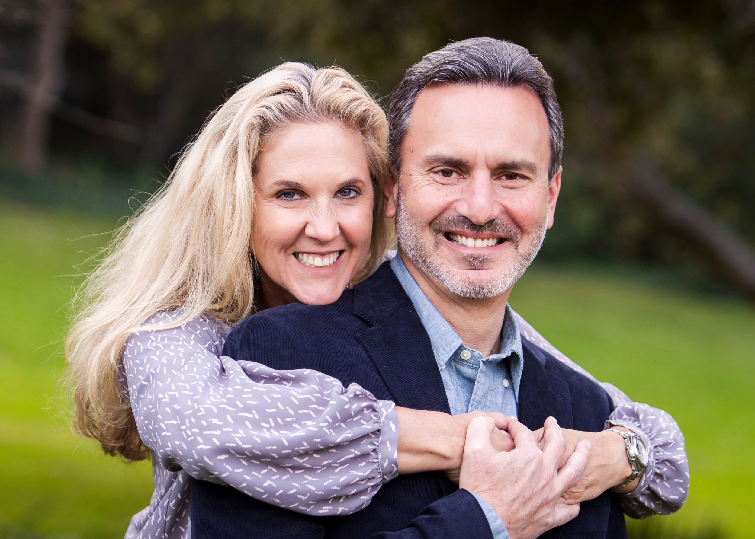 Ray and Kristen Mueller embrace outdoors with a green, blurred background.