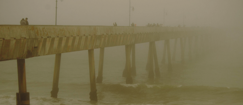 A foggy pier extending over the ocean with people walking and standing on it.