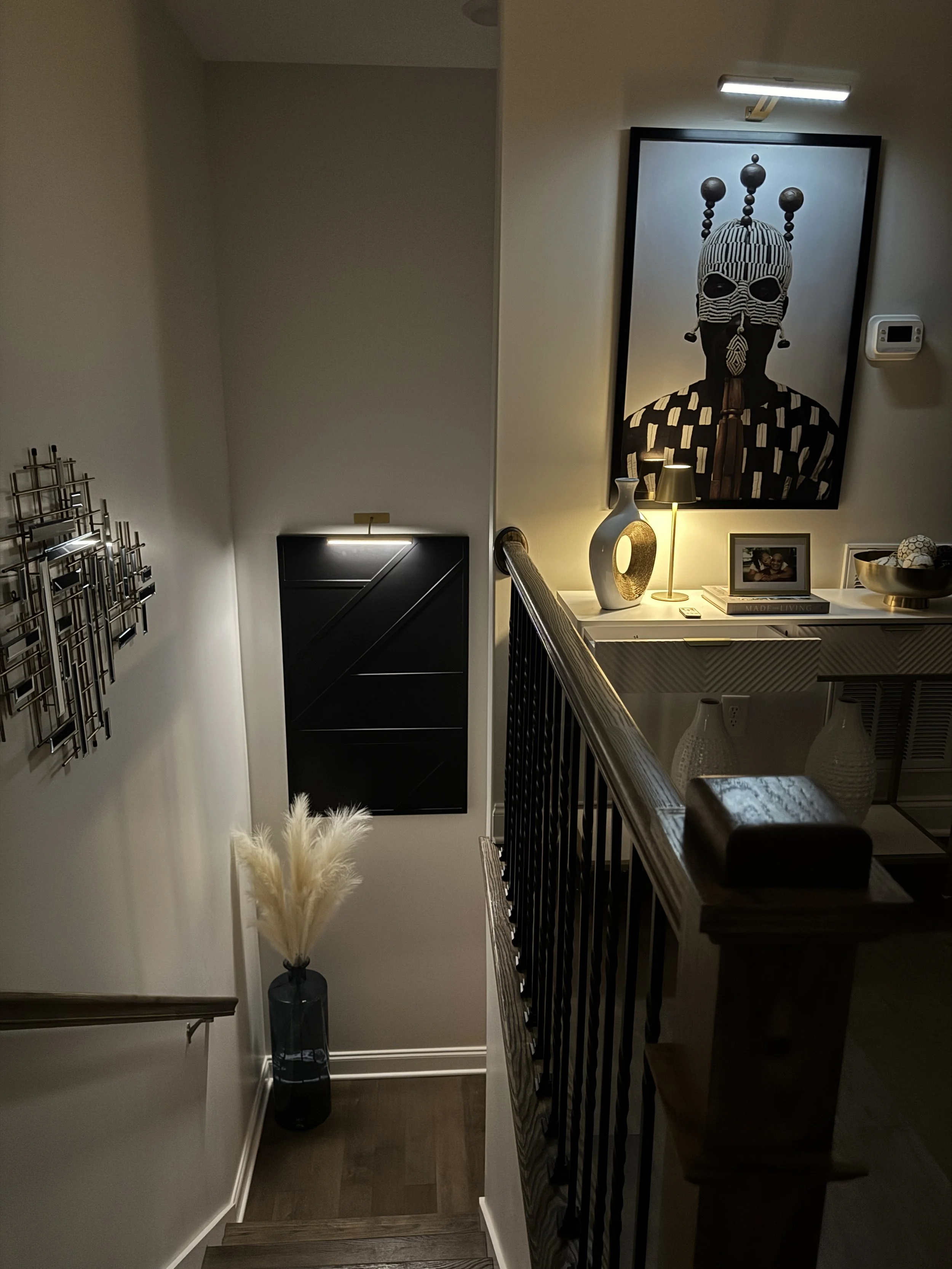 Interior view of a hallway with stairs, black wall art, decorative vase with pampas grass, and a side table decorated with a framed photo, sculpture, lamp, and bowl filled with decorative items in Oak Ridge home.