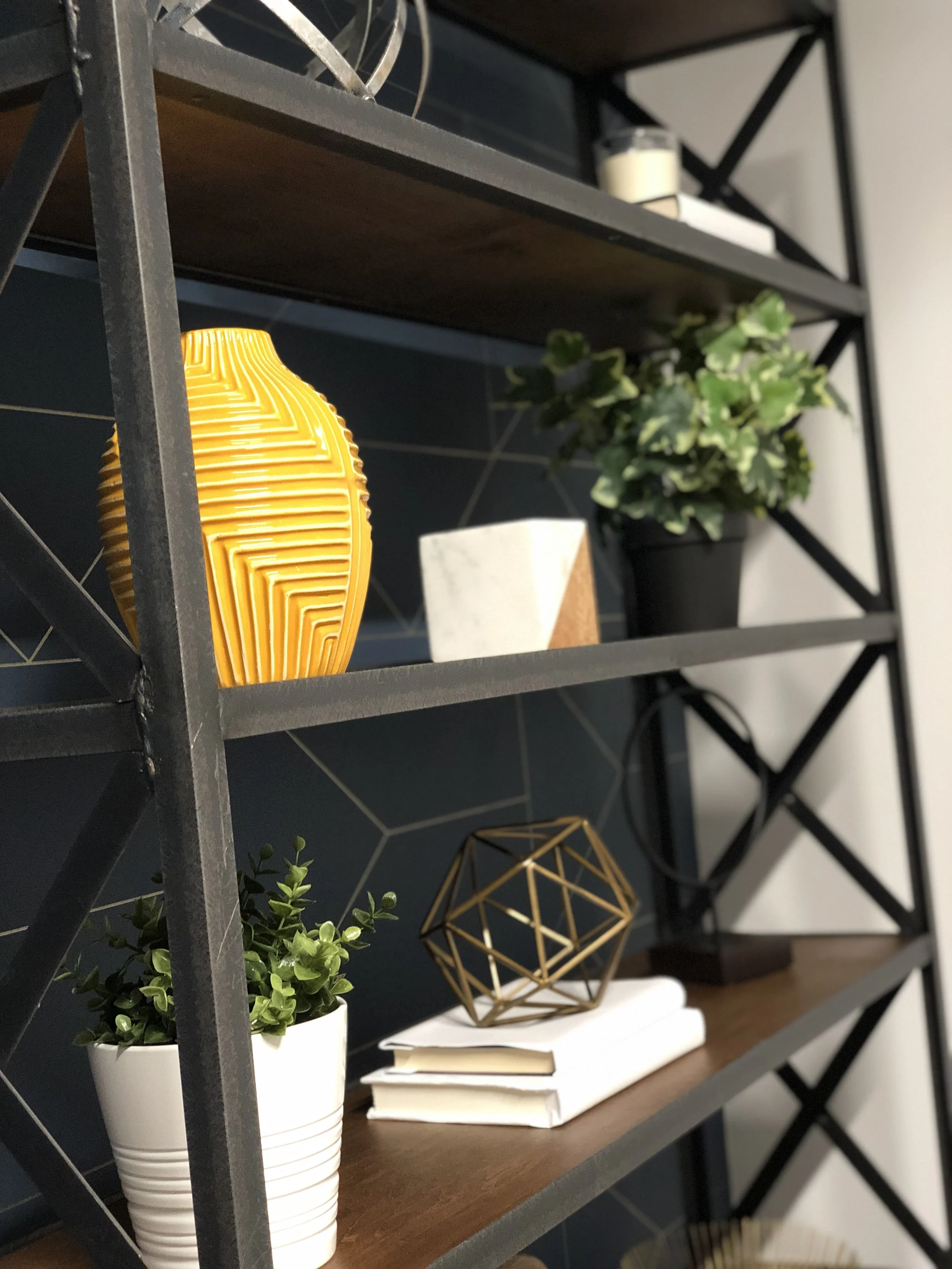 Shelving unit with decorative items including green plants in white pots, a yellow textured vase, a geometric gold sculpture, stacks of books, and a small candle holder. The background features a dark tiled wall in in Farragut.