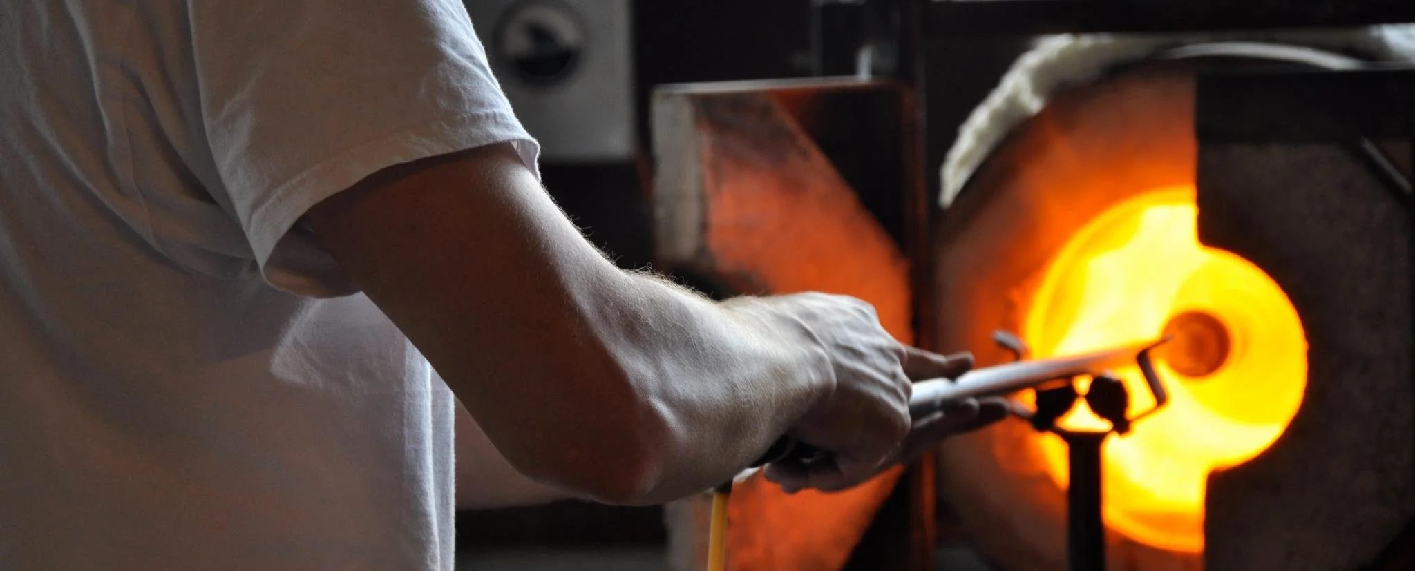 A person is holding a glowing metal blowpipe in a glassblowing furnace, shaping hot glass.