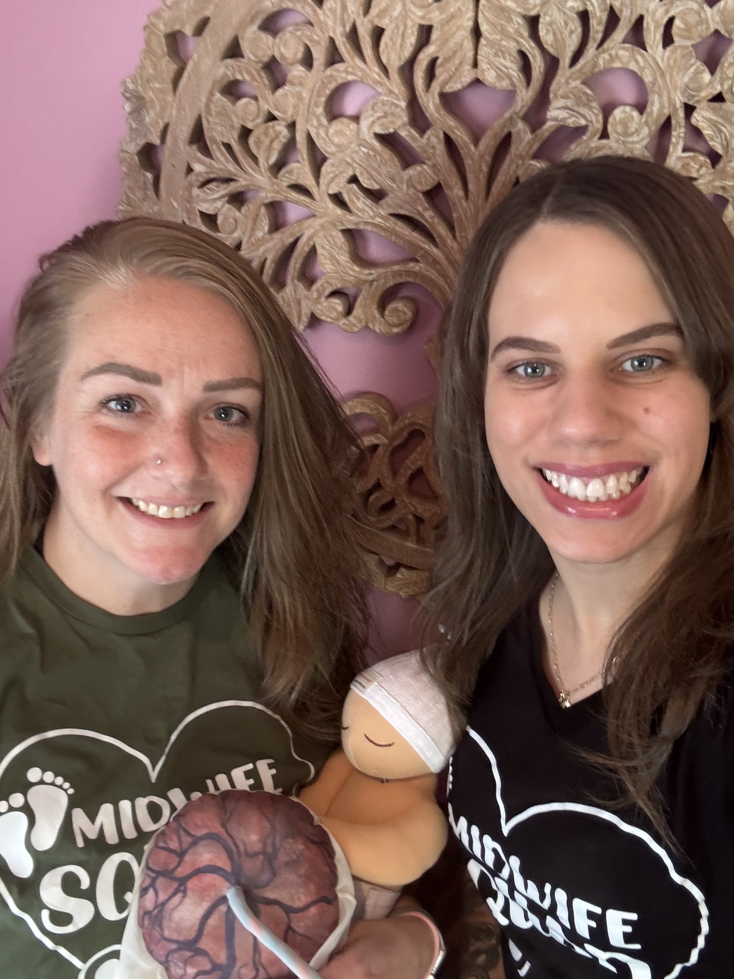 Two women smiling, holding a stuffed toy and a model of a brain, standing in front of a decorative wooden wall hanging. Both wear black shirts with a heart and brain graphic and text.