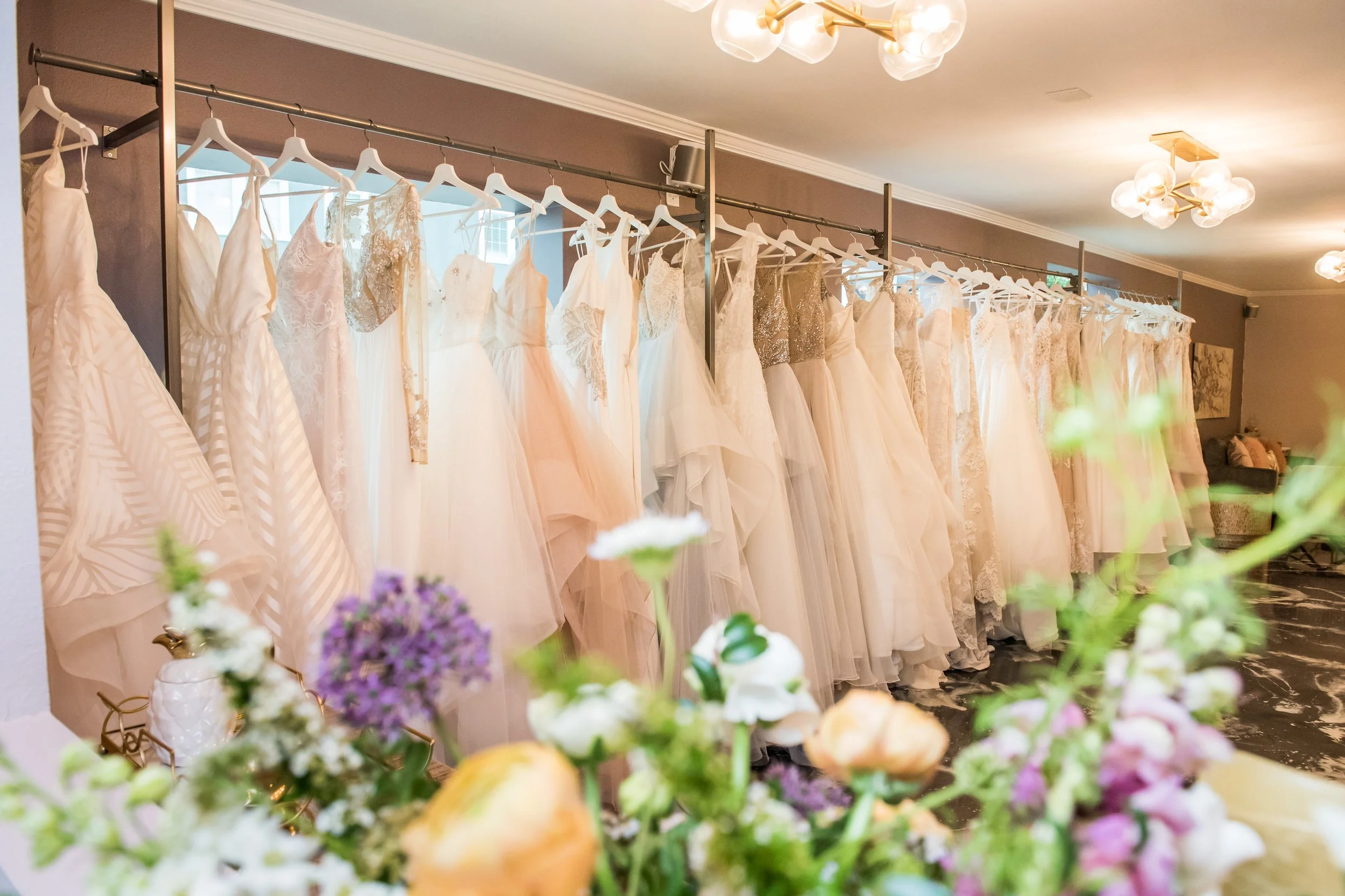 A row of white and cream-colored wedding dresses hanging on a rack in a bridal boutique with floral arrangements in the foreground.
