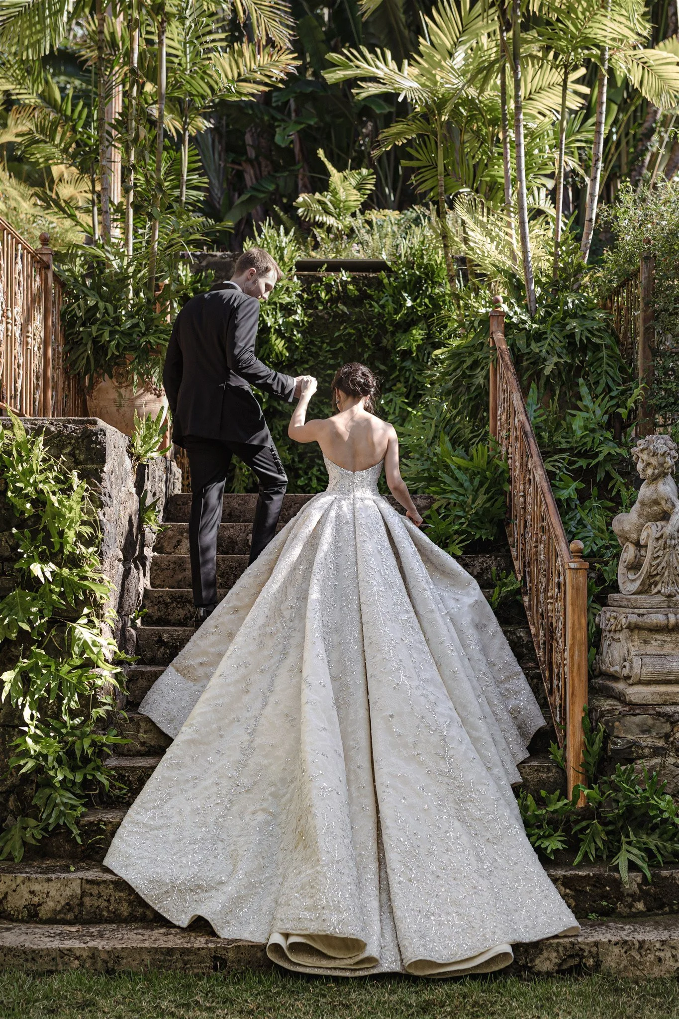 A bride in a strapless, ornate wedding gown with a long, flowing train and a groom in a black suit, holding hands and walking up stone stairs surrounded by lush greenery and decorative stone statues.