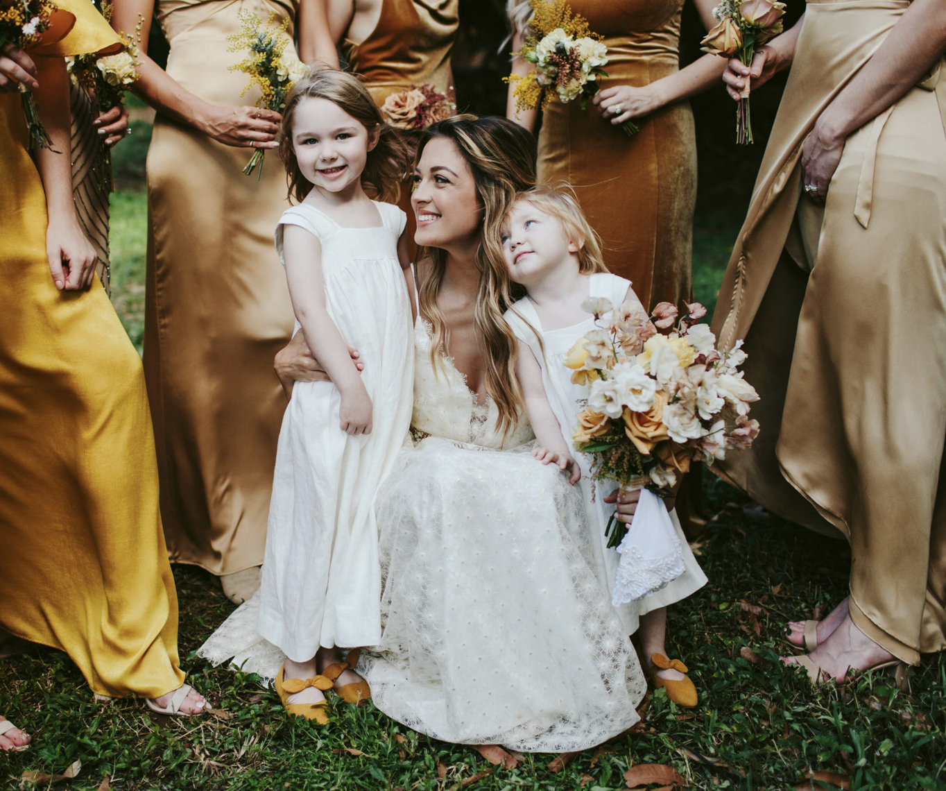 A woman with long, wavy hair in a white lace dress sitting on grass surrounded by women in gold dresses holding bouquets, with two young girls in white dresses, one hugging her and the other looking up at her, at a wedding or similar celebration.
