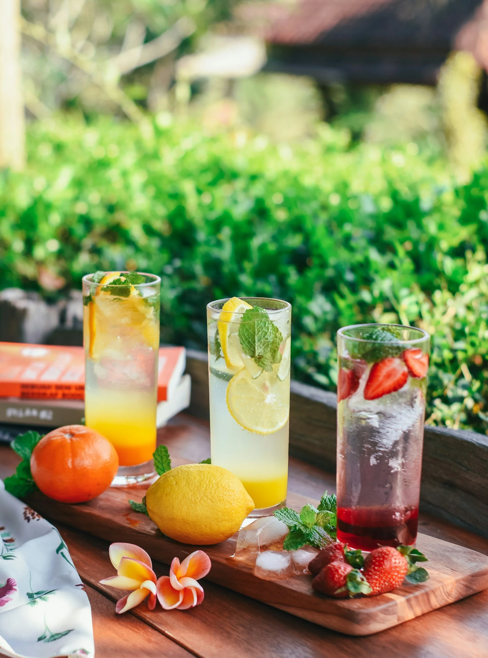 Three glasses of fruit-infused beverages on a wooden serving board, garnished with lemon, mint, and strawberries, with fresh fruit and flowers beside them, set outdoors on a sunny day.
