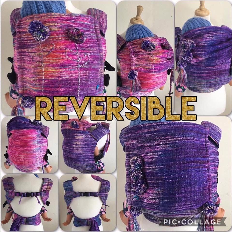 Colorful, knit baby carrier with a reversible design showing pink and purple tones, adorned with small pom-pom decorations, displayed on a mannequin with front and back views.