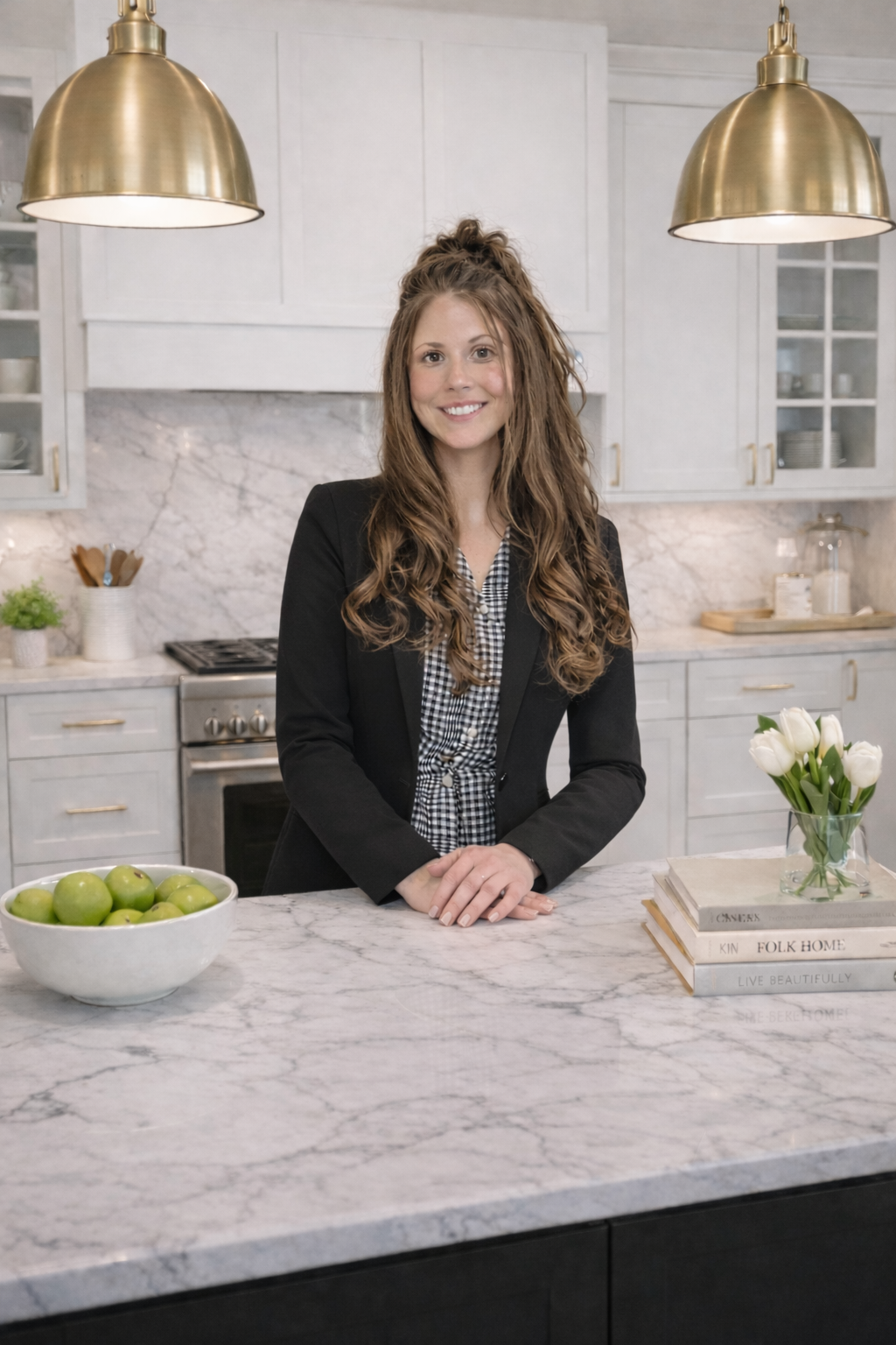 A woman with long, curly brown hair, wearing a black blazer and a checkered shirt, stands in a modern kitchen with white cabinets, a marble island, and gold pendant lights. A bowl of green apples and a stack of books with a glass vase of white tulips are on the countertop.
