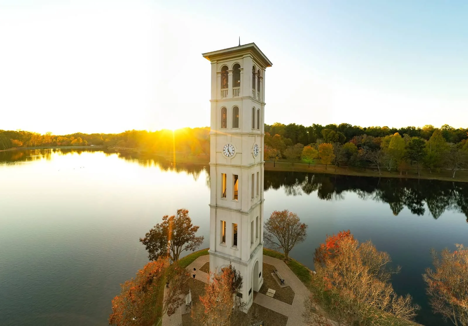 A tall white clock tower situated on a small island in a body of water, with trees showing fall colors around it, during sunset with the sun near the horizon.