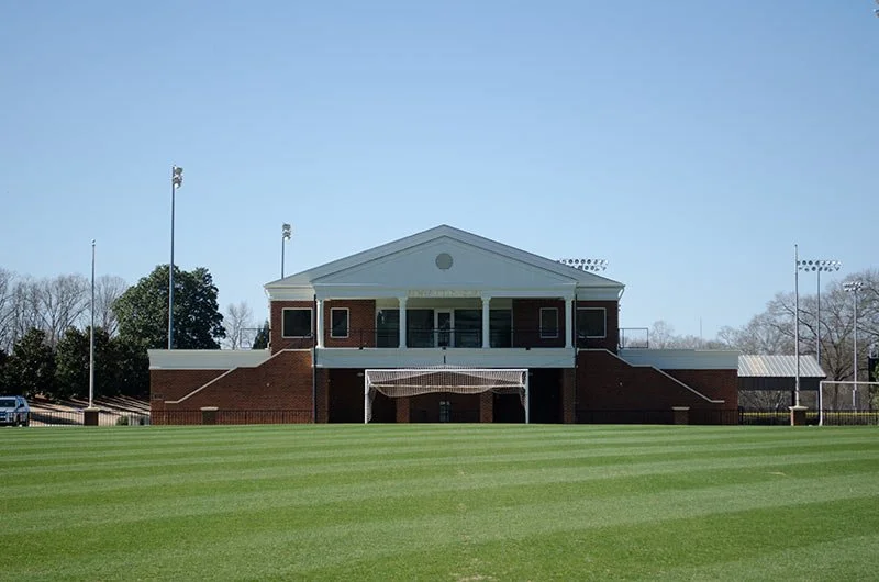 A sports stadium building with a football field in front and stadium lights visible in the background.