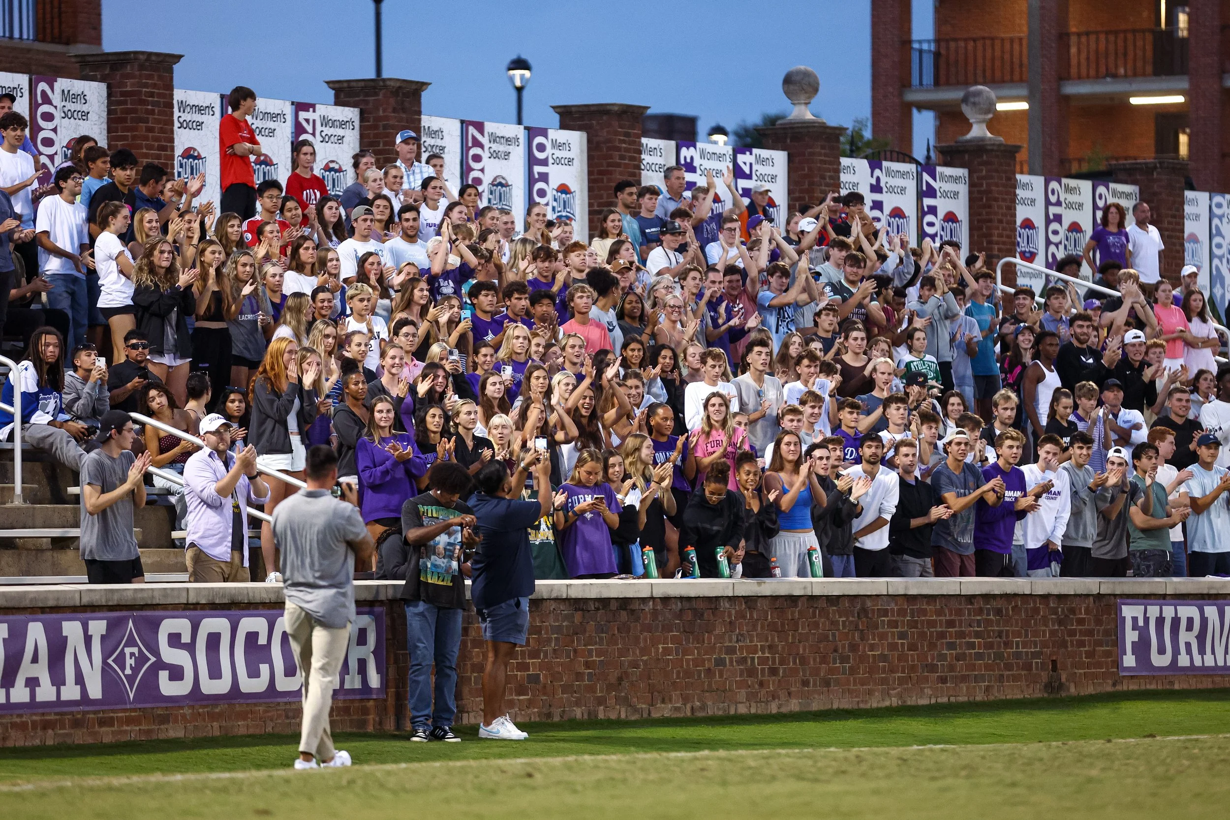 Crowd of spectators at a soccer game, standing and cheering in stadium stands with banners reading 'Women's Soccer' and 'Men's Soccer', some taking photos or videos.