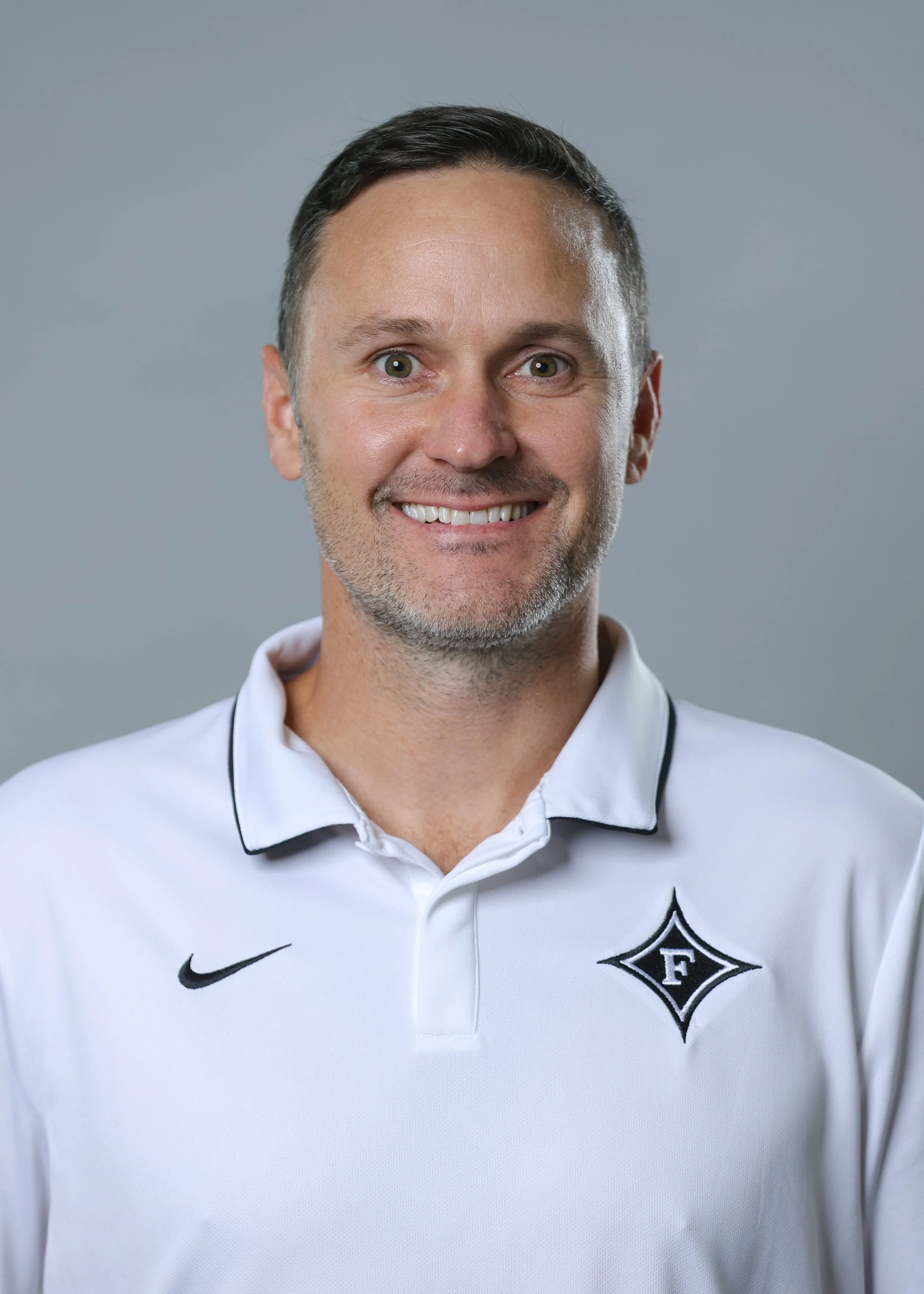 A smiling man with short dark hair, a five o'clock shadow, wearing a white sports polo shirt with black accents and a logo on the chest.