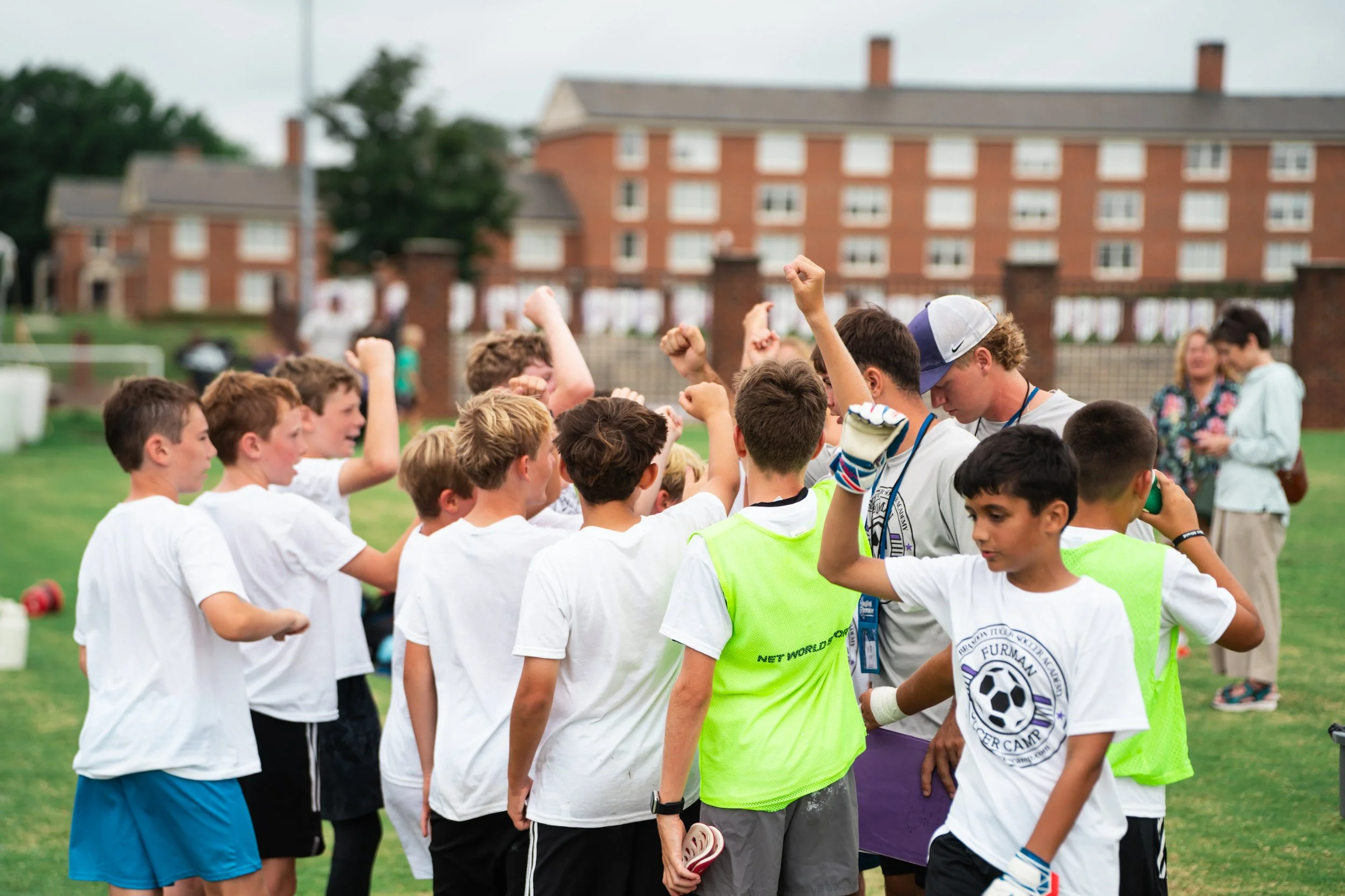 Crowd of people at an outdoor event, some clapping and cheering. A young man in a purple shirt and sunglasses holds a white sign that says "Let's Go Dinos".