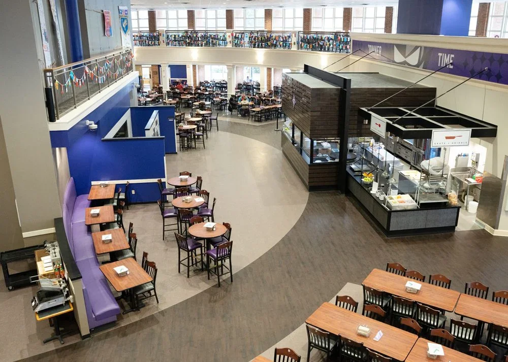 Interior of a cafeteria or food court area with tables and chairs, a service counter, and a second-floor balcony with more seating.