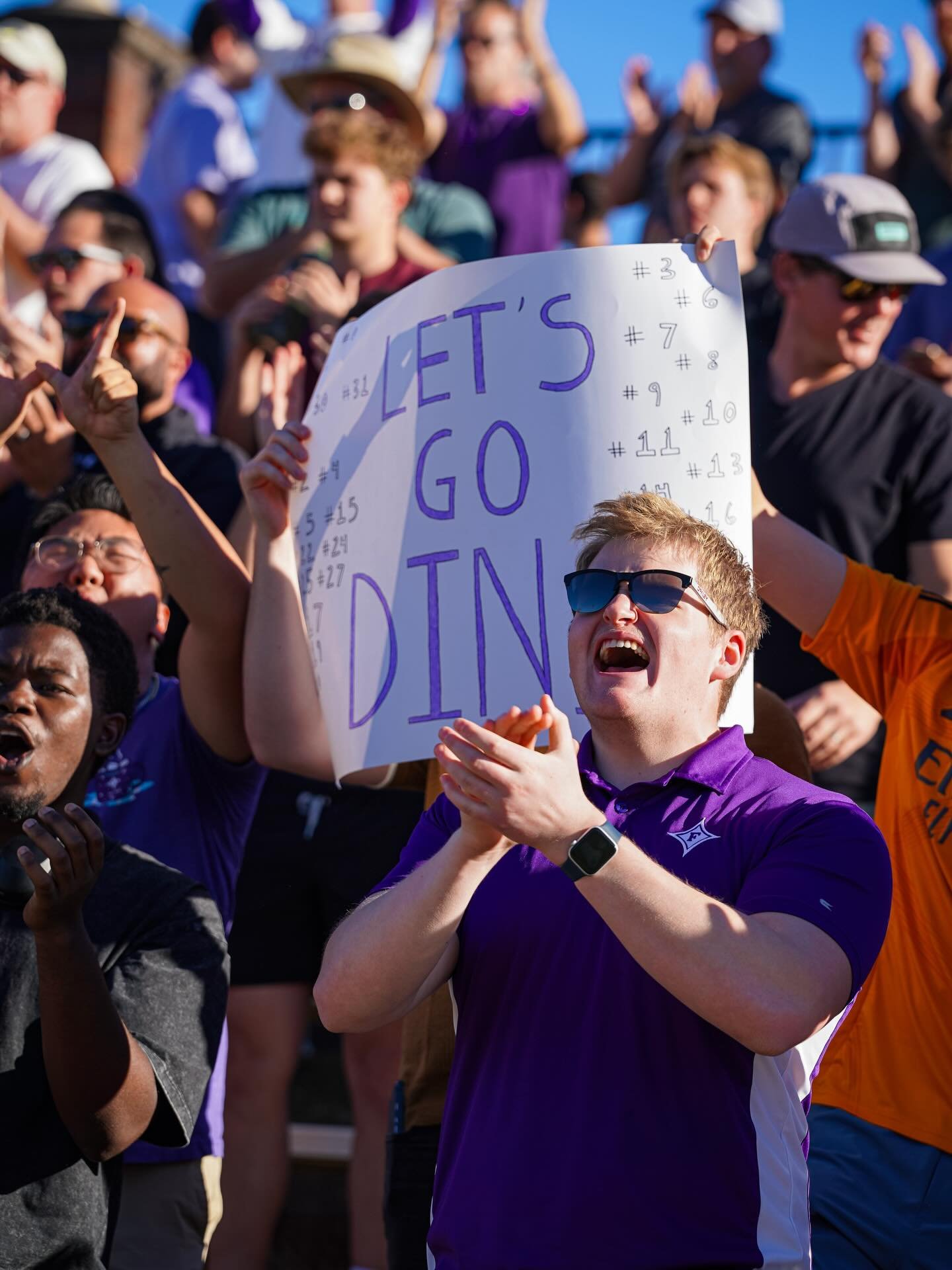 Crowd of people at an outdoor event, some clapping and cheering. A young man in a purple shirt and sunglasses holds a white sign that says "Let's Go Dinos".